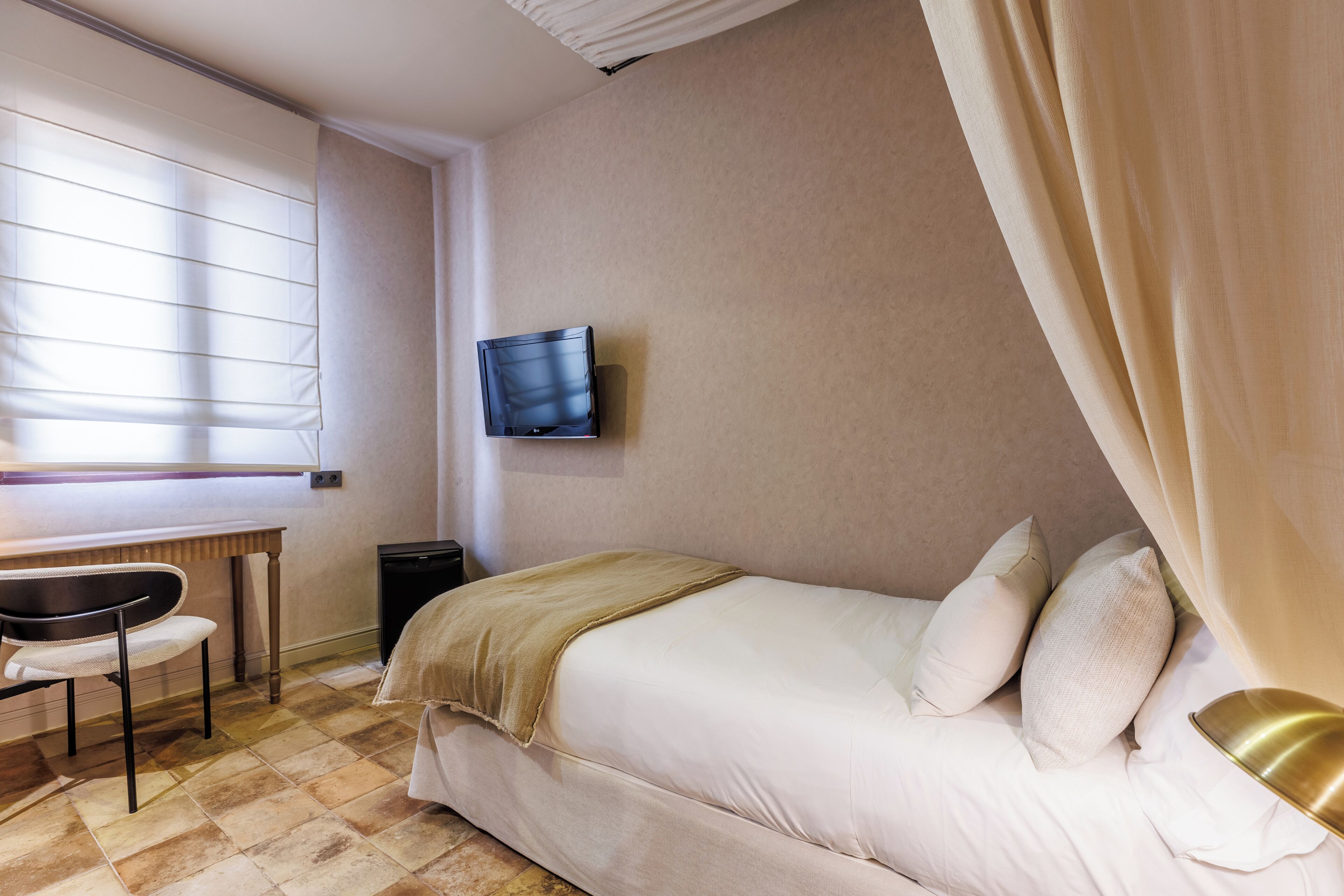 A hotel room with a bed, a desk, a chair, a television mounted on the wall, and a window with blinds.