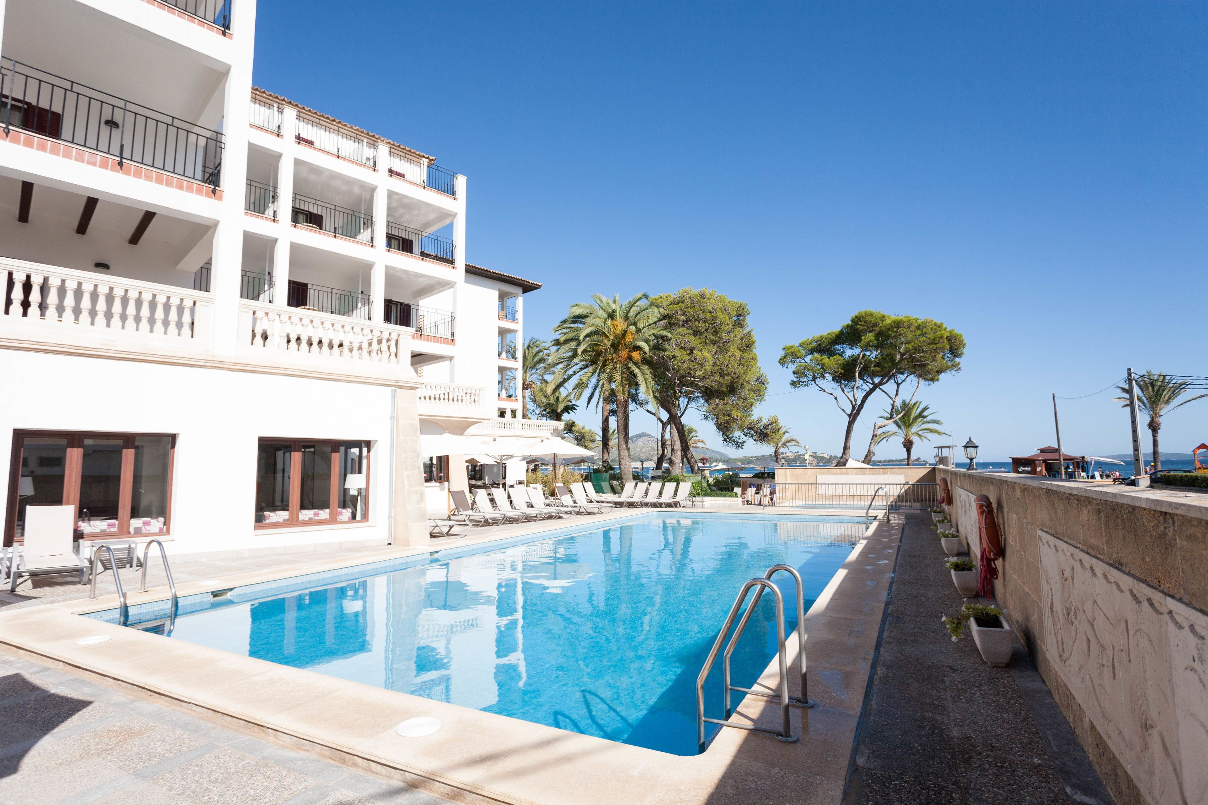 A swimming pool with lounge chairs and umbrellas, a white building with balconies, and palm trees.