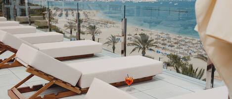 A beachside hotel balcony with sun loungers and a glass railing overlooking the beach and mountains.