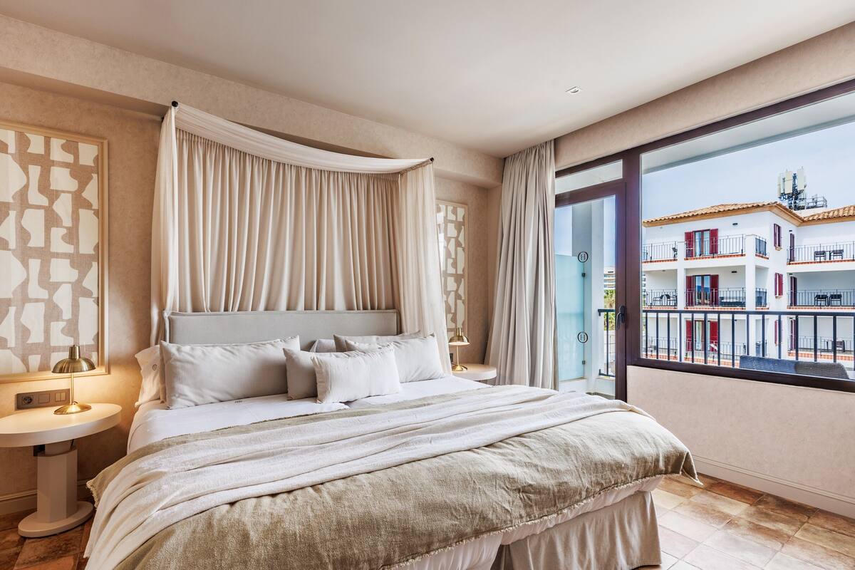 Quarto com uma cama grande, mesa de cabeceira, luminária e vista para um edifício através de uma janela ampla.