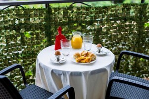 Mesa redonda com toalha branca, jarra de suco de laranja, dois copos, prato com croissants, porta-guardanapos vermelho e um ovo pequeno numa colher.