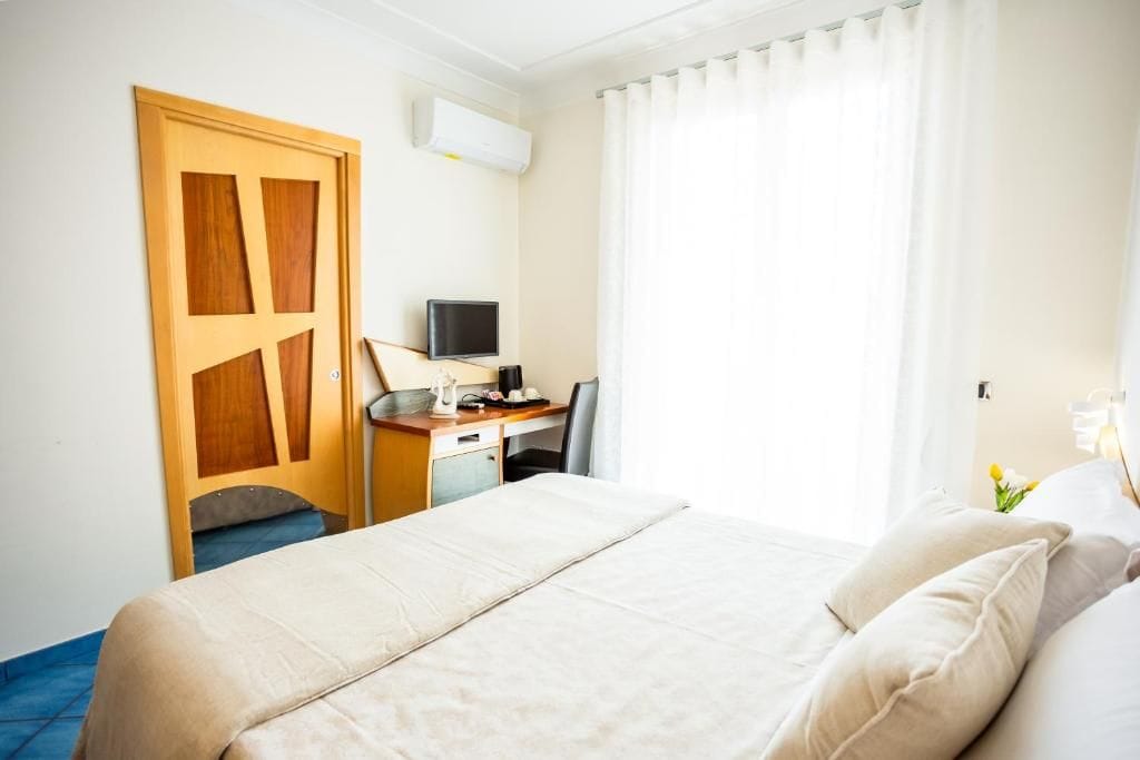 A hotel room with a bed, a desk with a computer, a chair, and a window with curtains.