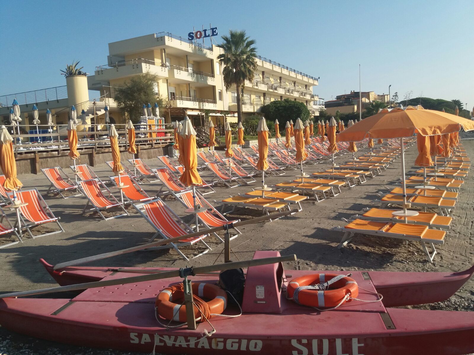 A beach with rows of orange and white striped lounge chairs and yellow umbrellas. In the background, there is a building with the sign "SOLE".