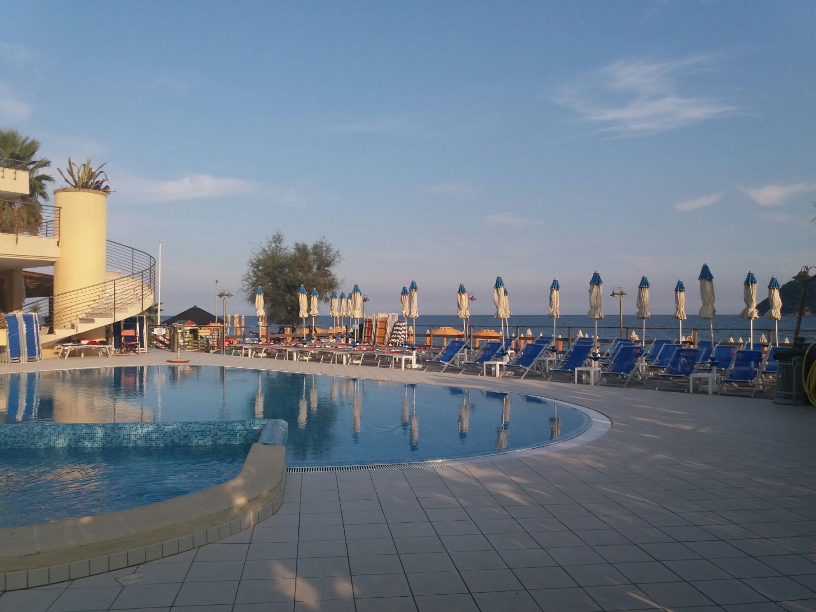 A pool area with blue lounge chairs and umbrellas, a clear sky, and a view of the sea.