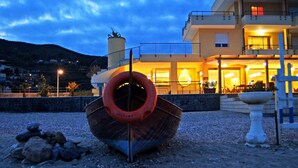 A boat with a life ring on a pebbled beach, a building with balconies and lights, and a mountainous backdrop. - Residence Sole (Albenga)