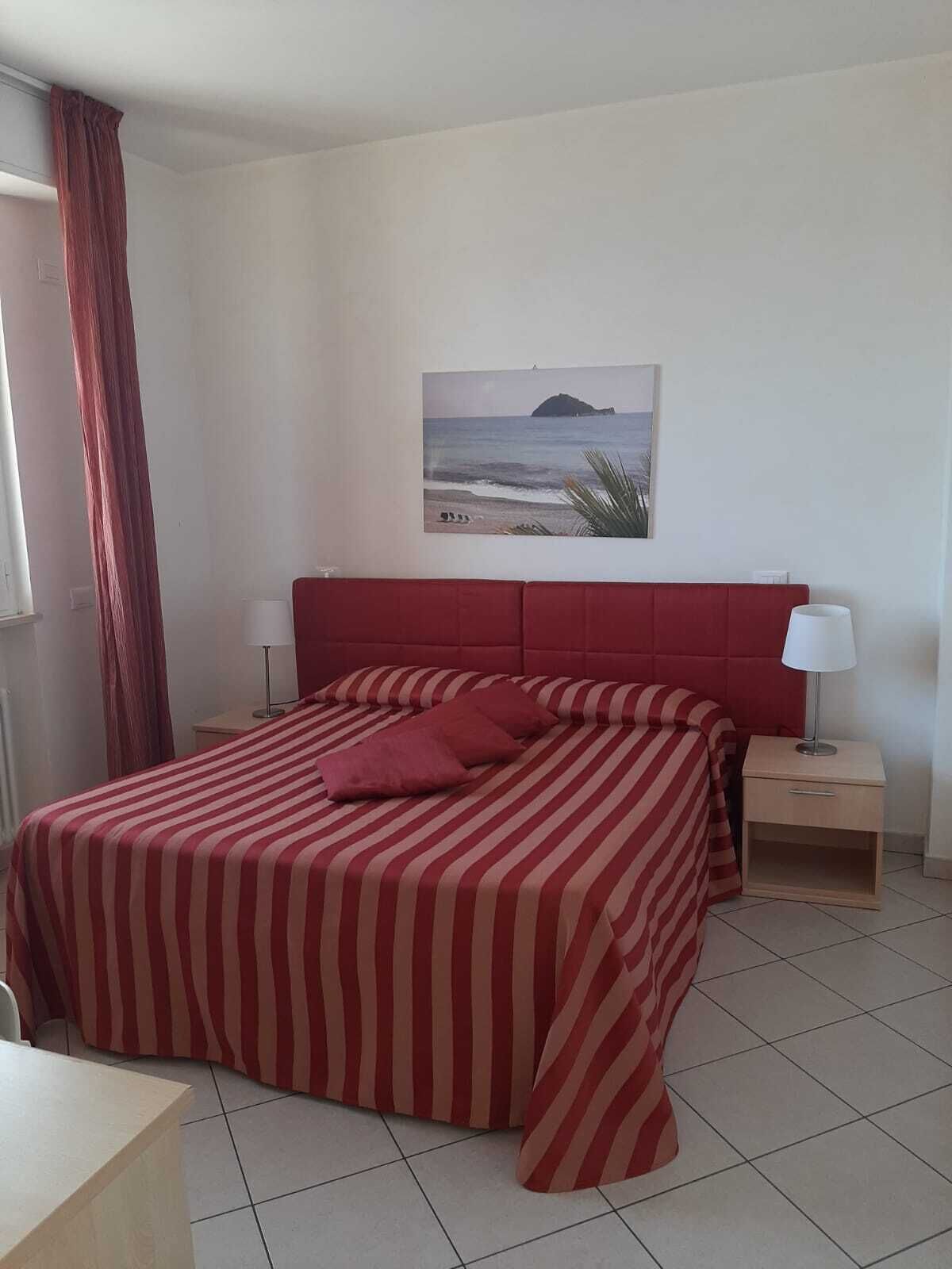 A hotel room with a bed, a nightstand, a lamp, a window with curtains, and a wall-mounted picture of a beach.