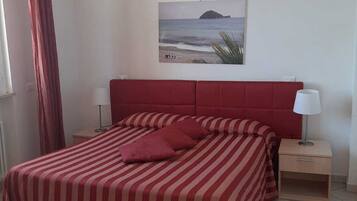 A hotel room with a bed, a nightstand, a lamp, a window with curtains, and a wall-mounted picture of a beach.