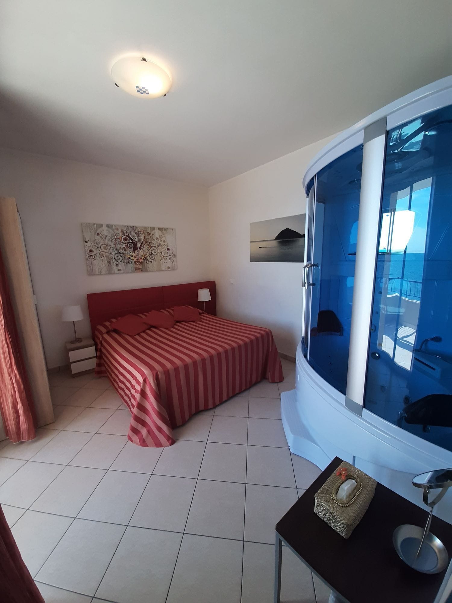 A bedroom with a bed, a nightstand, a window with a view, and a small table with a lamp.