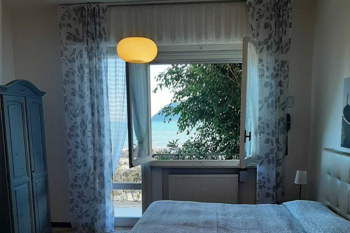 A bedroom with a bed, a desk, a chair, and a view of the sea through a window.