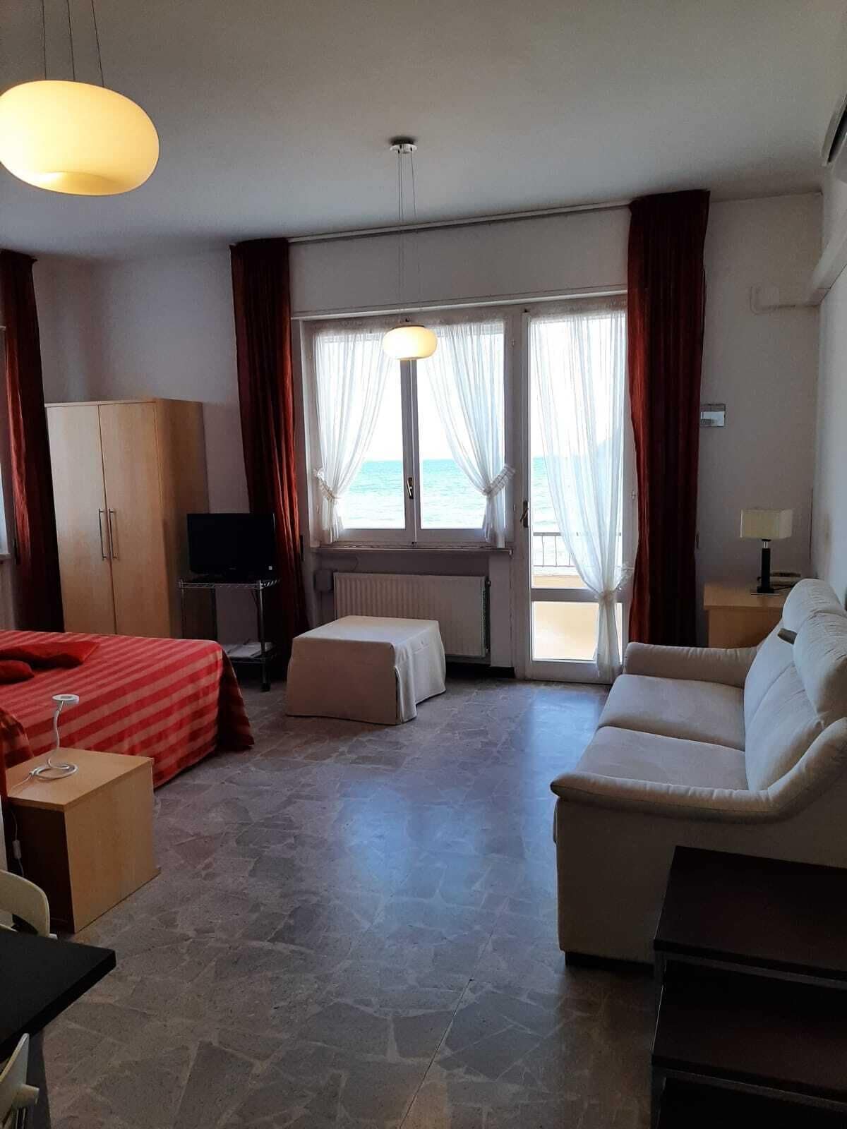 Habitación de hotel con cama, sofá, televisión, armario y vista al mar a través de amplias ventanas.