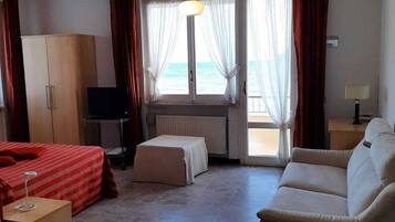 Habitación de hotel con cama, sofá, televisión, armario y vista al mar a través de amplias ventanas.