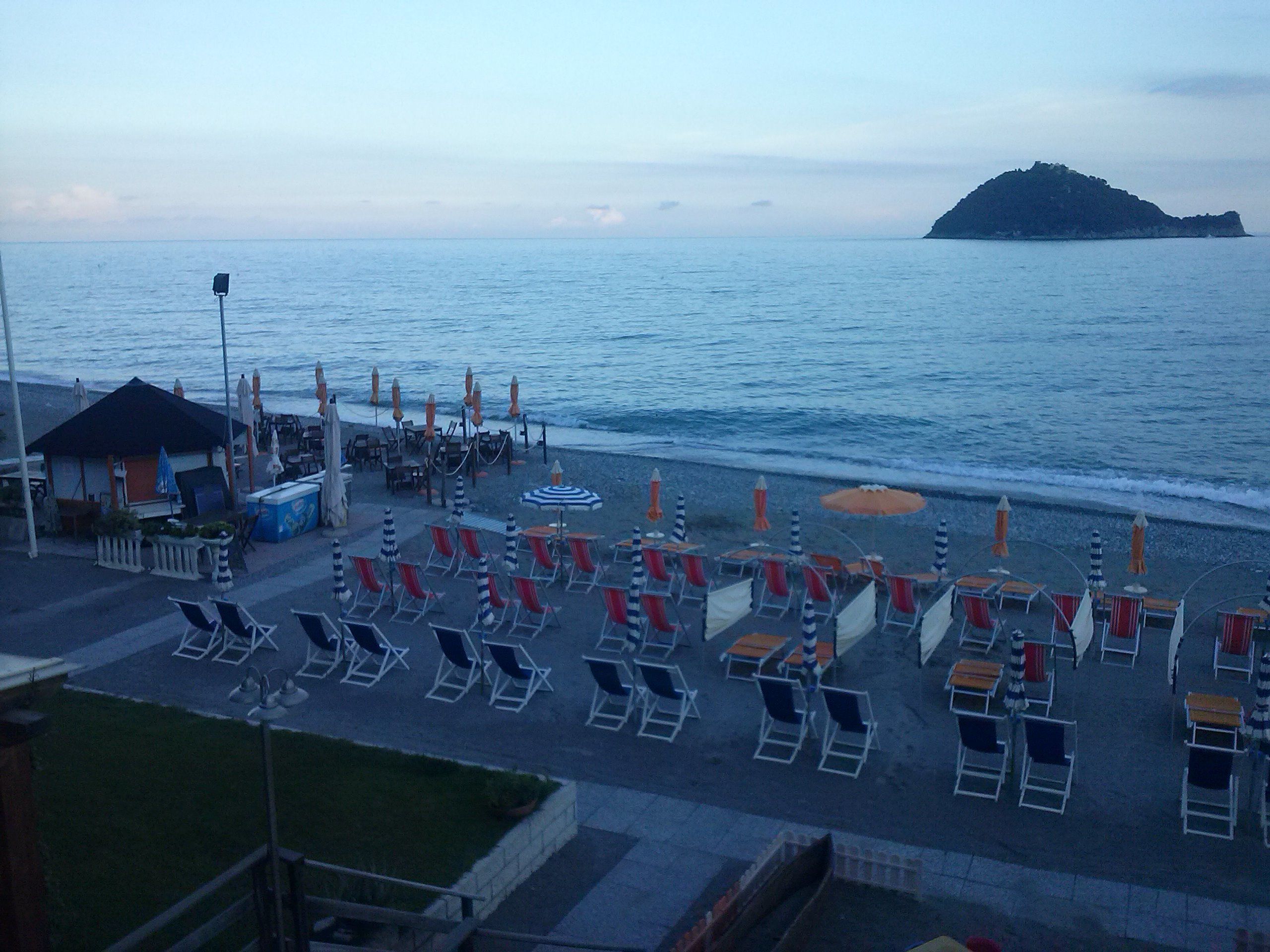 A beachside seating area with rows of sun loungers and umbrellas, a small building, and a view of the sea and a distant island.