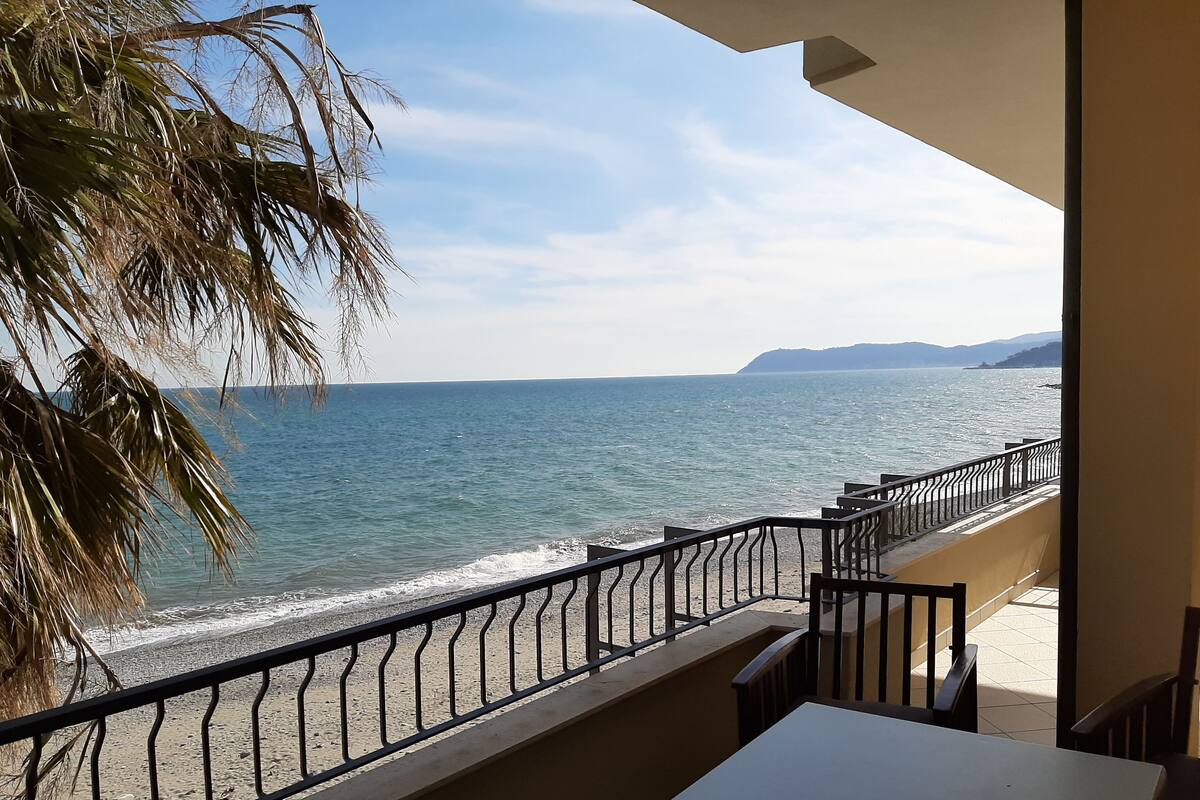 A balcony with a view of the sea, a palm tree, and a tiled floor.