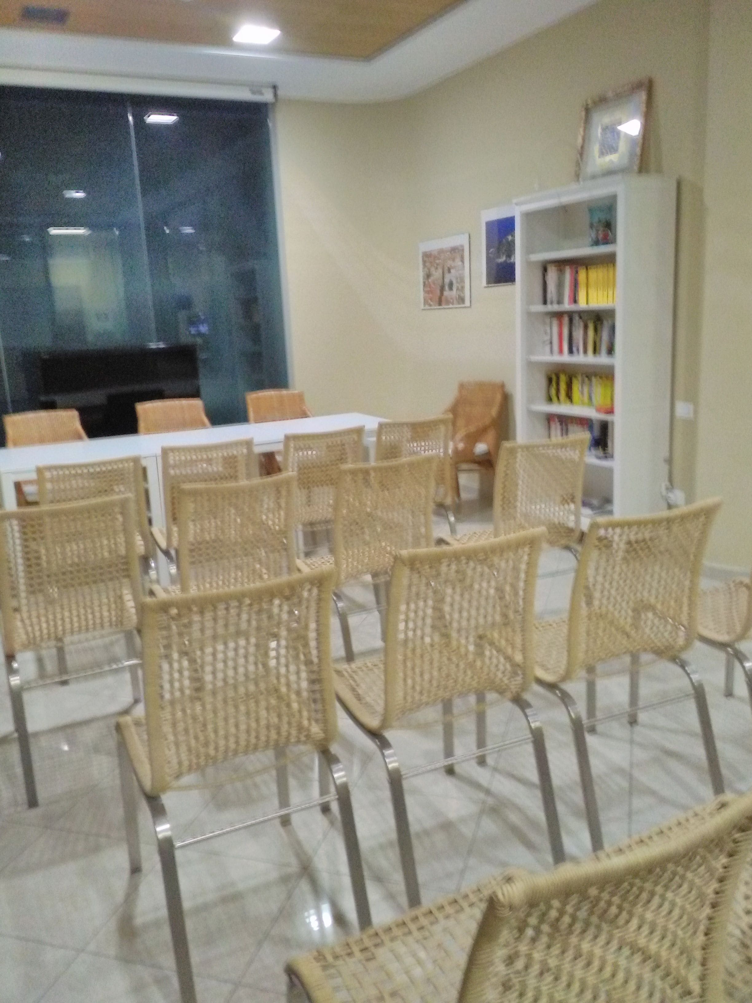 Un salón de conferencias con sillas de mimbre, una mesa blanca y una estantería llena de libros.