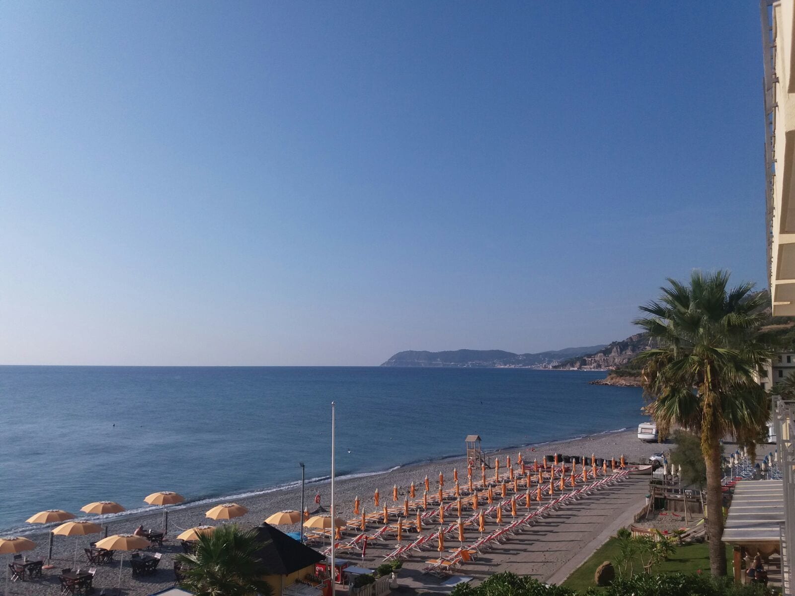 Una playa con hileras de sombrillas y sillas de descanso, un cielo azul despejado, y vistas al mar y colinas distantes.