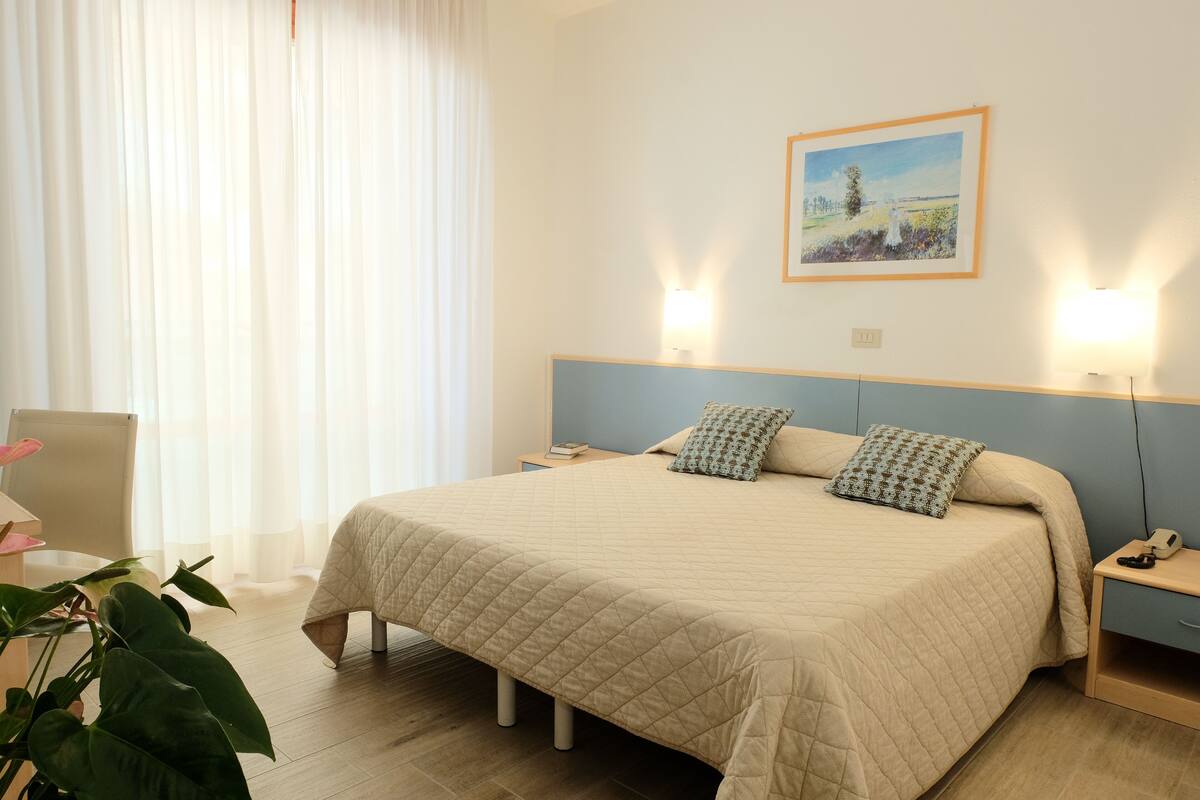 A neatly made bed with a quilted bedspread, two pillows, bedside tables with lamps, a framed picture on the wall, and a potted plant.