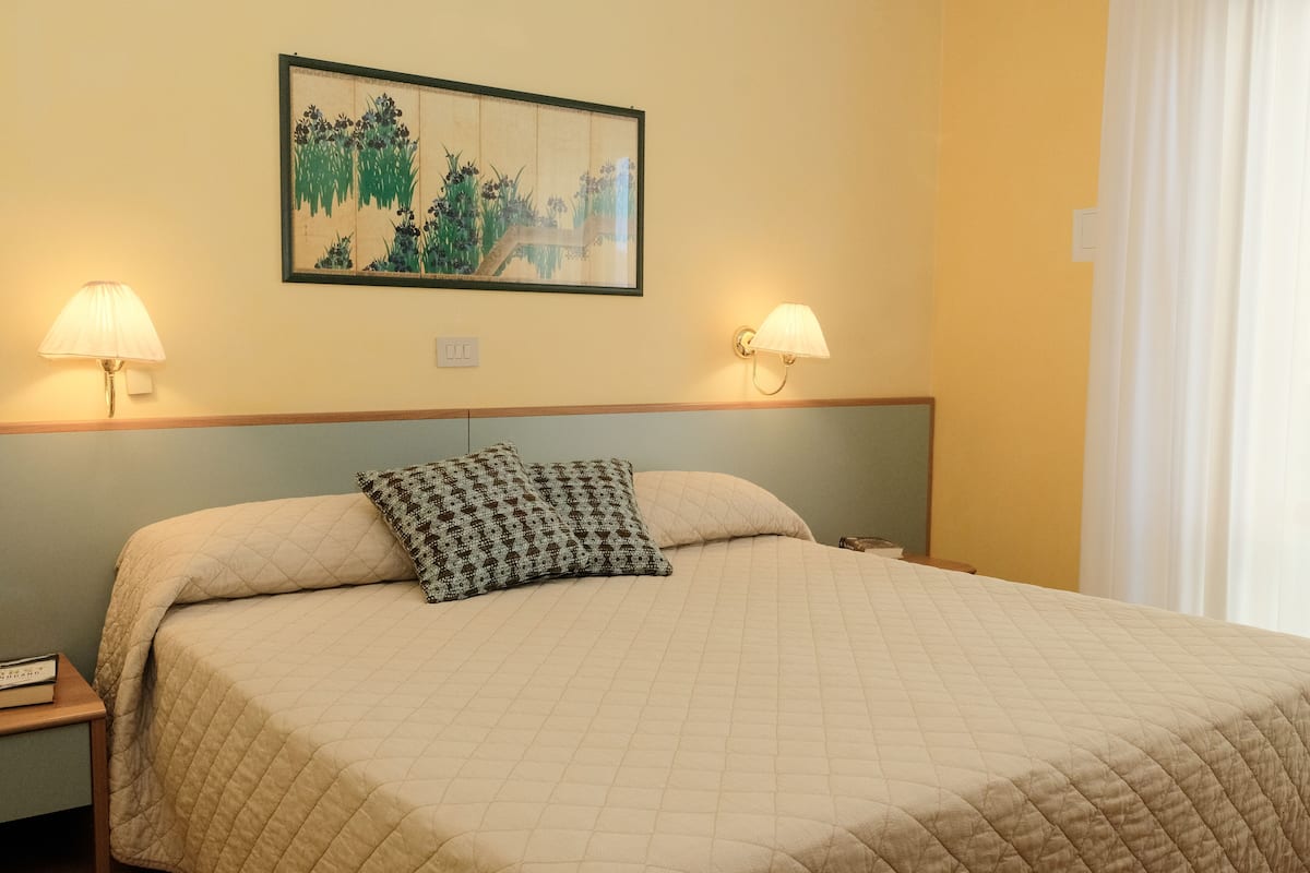 A neatly made bed with a quilted bedspread and two pillows, a wall-mounted painting, and two bedside lamps.