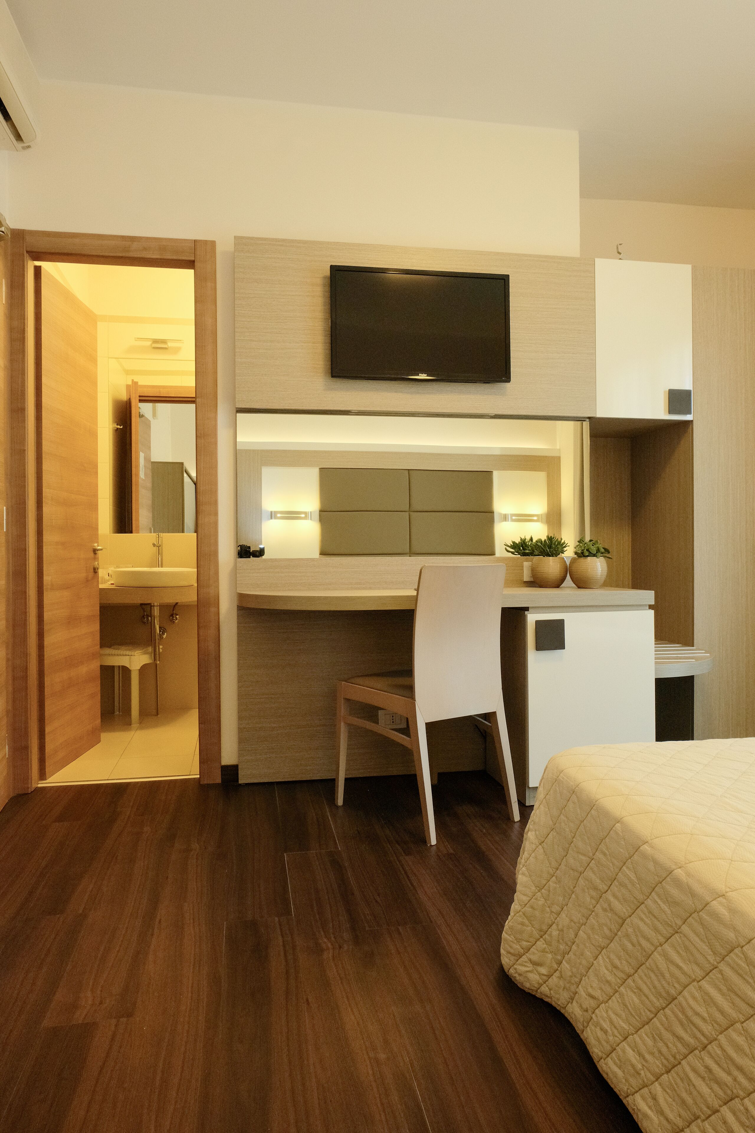 Una habitación de hotel con piso de madera, una cama con colcha color beige, un escritorio con silla, un televisor montado sobre el escritorio, y una pequeña planta en maceta sobre el escritorio.