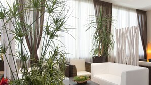 A modern living room with a white sofa, a wicker coffee table, and large potted plants. - Hotel Orizzonte (Jesolo)