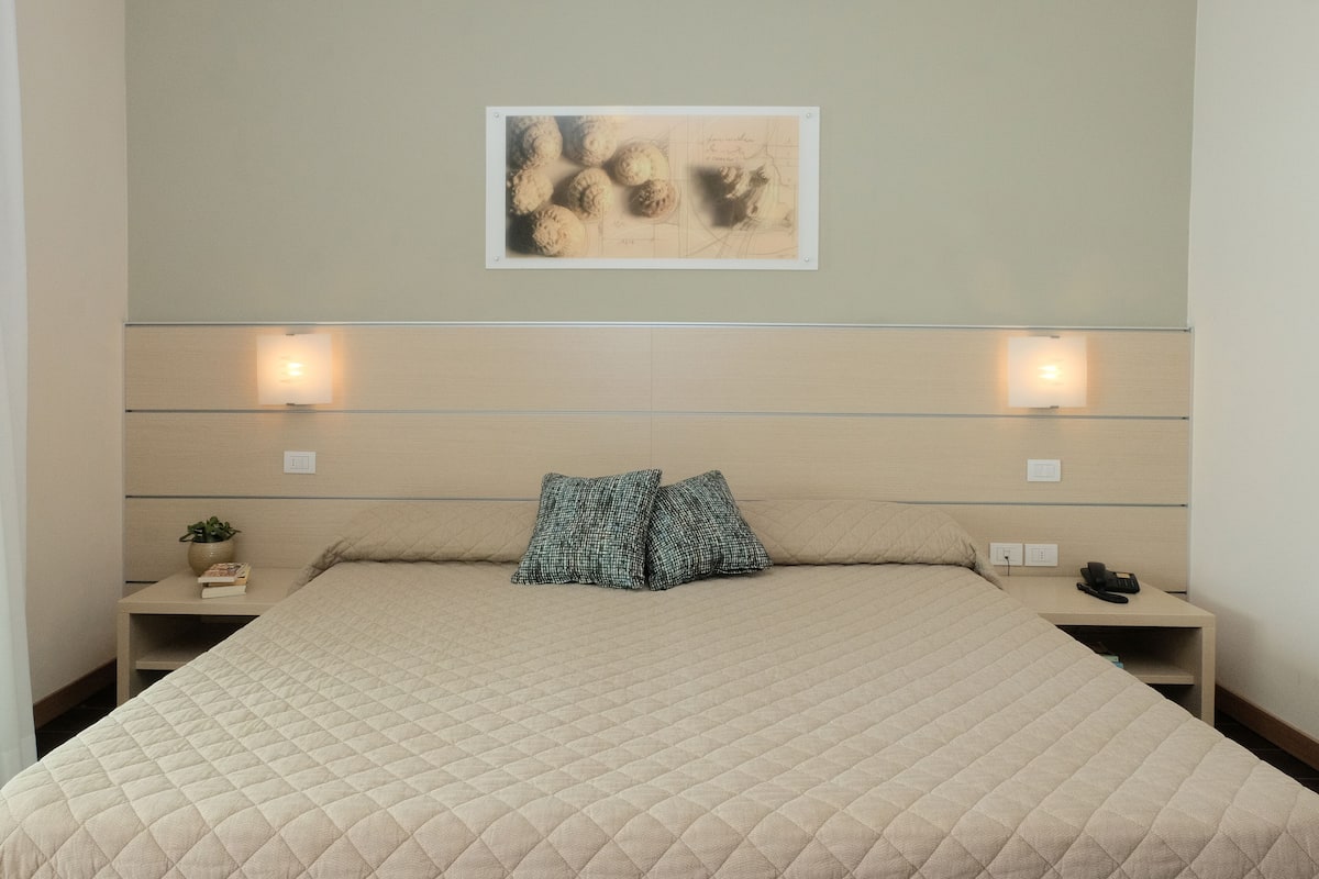 Ein ordentlich bezogenes Bett mit einer gesteppten Decke, zwei Nachttische mit Lampen, ein an der Wand befestigtes Kunstwerk und eine kleine Pflanze auf einem der Tische.