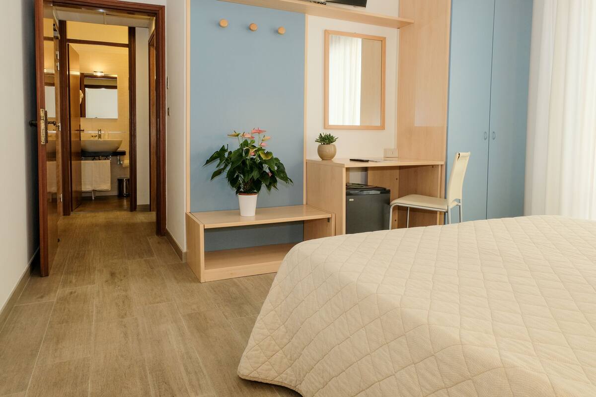 Ein Hotelzimmer mit Bett, Schreibtisch, Stuhl, einer kleinen Pflanze, einem Spiegel und einem durchgegangenen Tür sichtbaren Badezimmer.