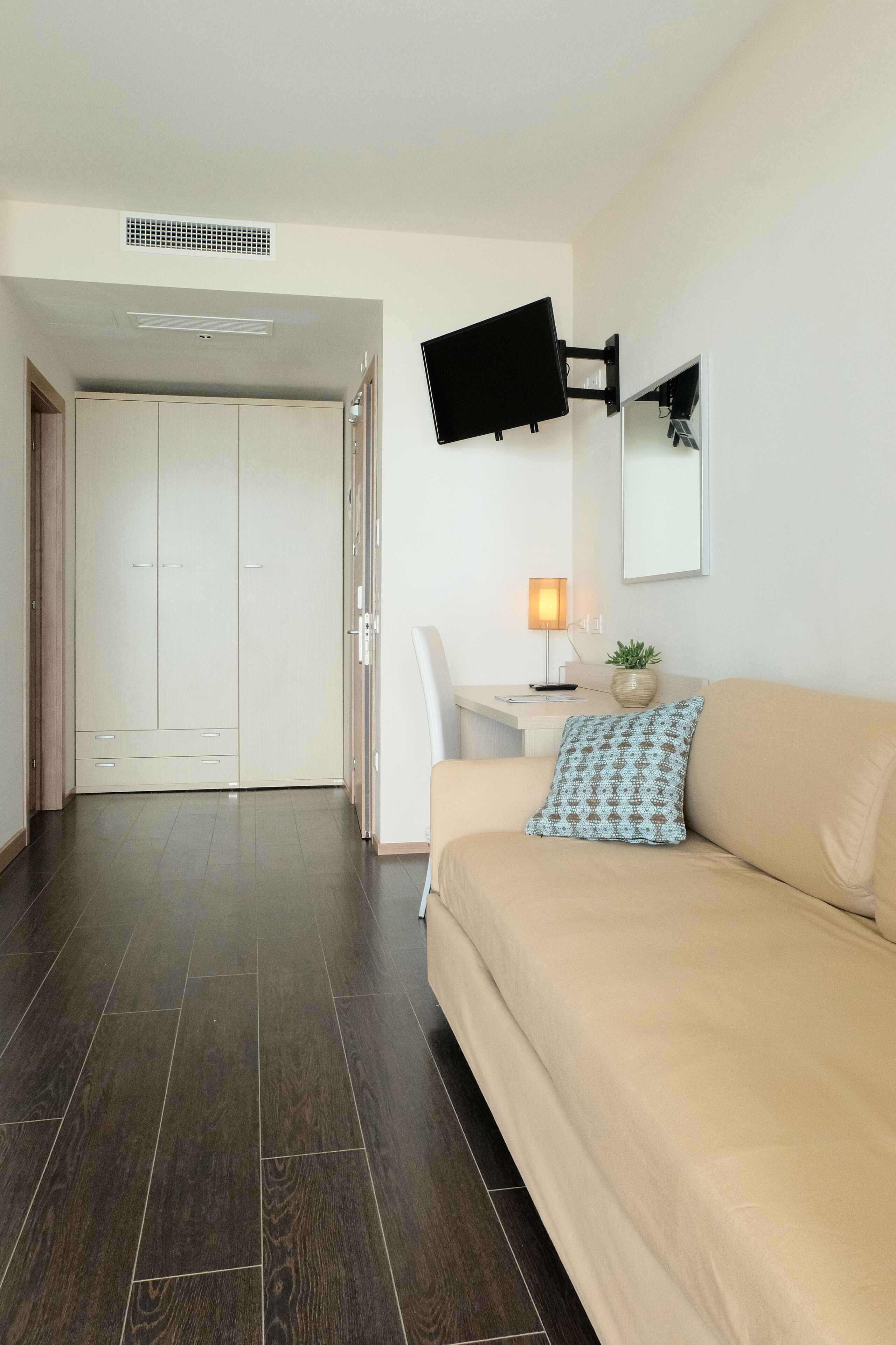 Een moderne hotelkamer met een beige bank, een bureau met een lamp, een flatscreen televisie aan de muur en een grote kast.