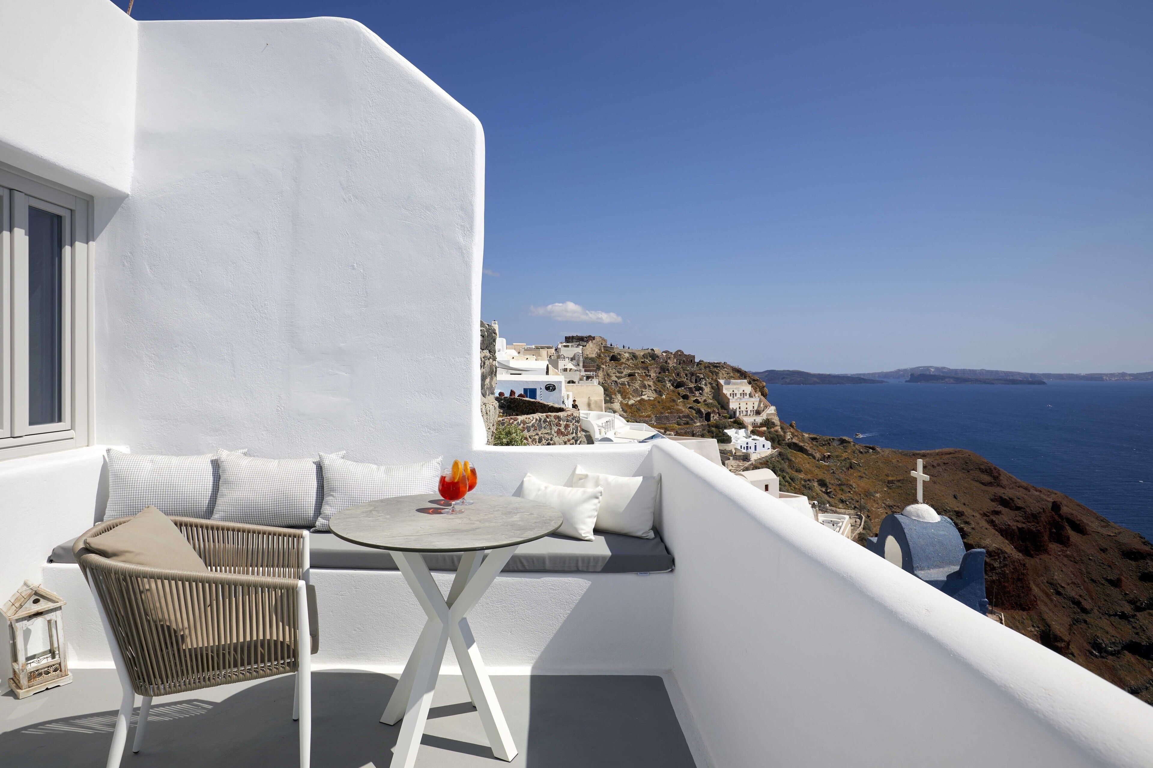 A balcony with a wicker chair, a small round table, and a glass of drink. The view includes a coastal landscape with white buildings and a blue sky.