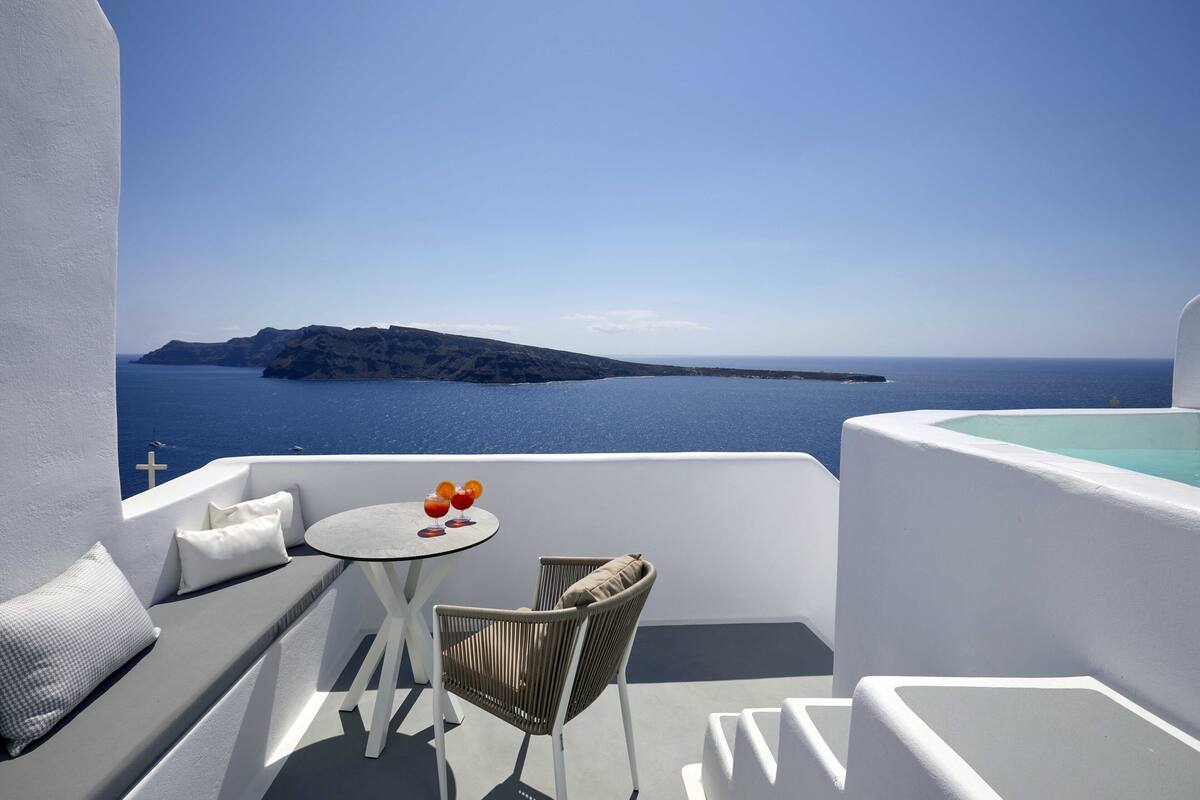 Een balkon met een witte inrichting, een ronde tafel met een stoel, en uitzicht op zee en een eiland in de verte.