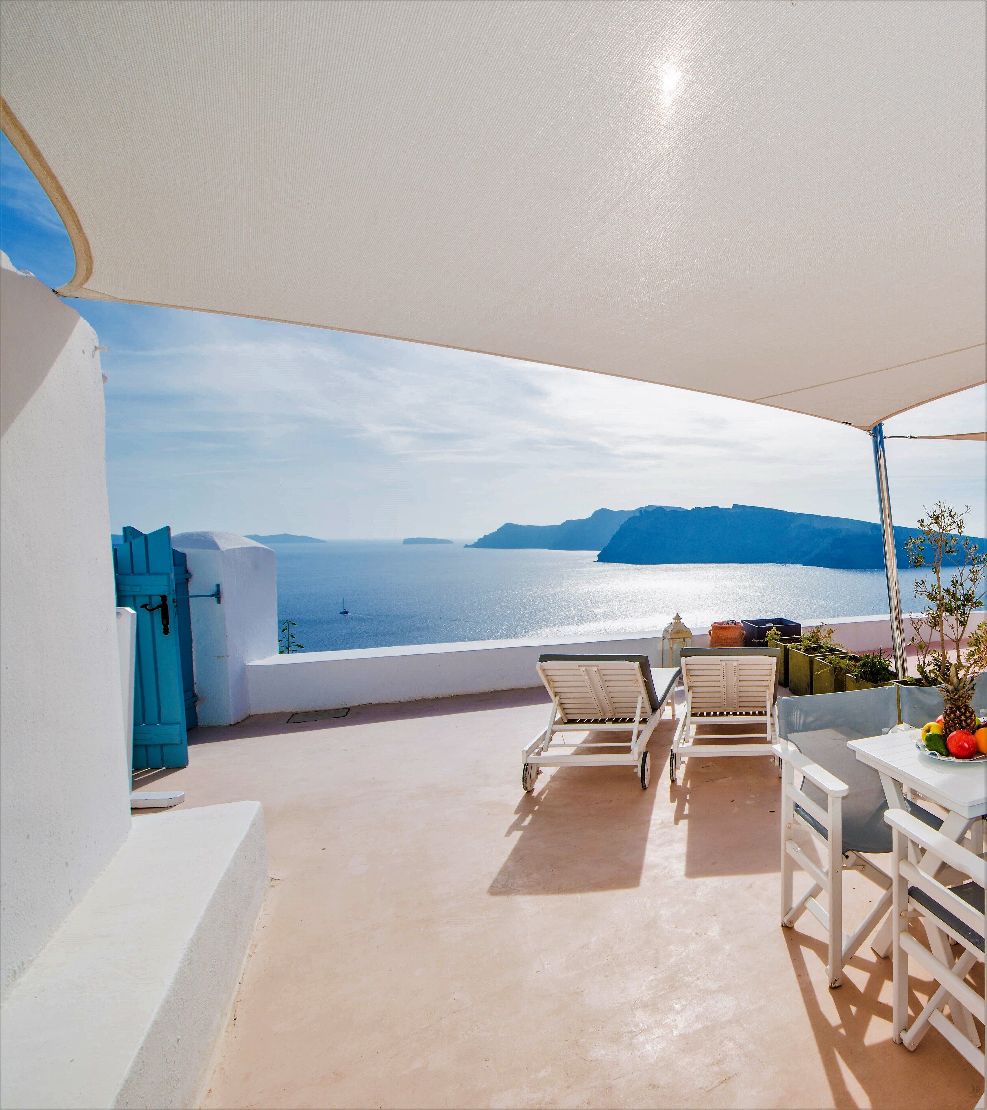 A terrace with white furniture, a table with fruit, and a view of the sea and mountains.