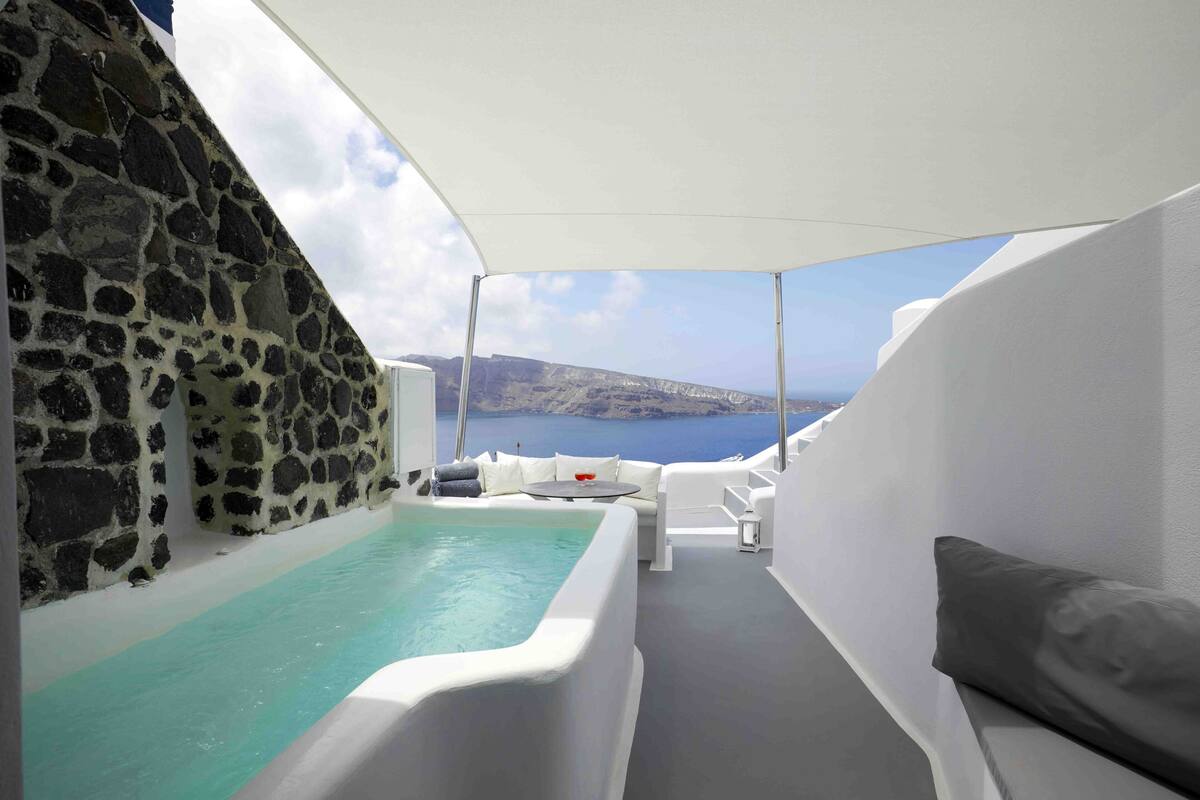 Een zonnig terras met een stenen muur, een jacuzzi, witte meubels en uitzicht op zee en een berg in de verte.