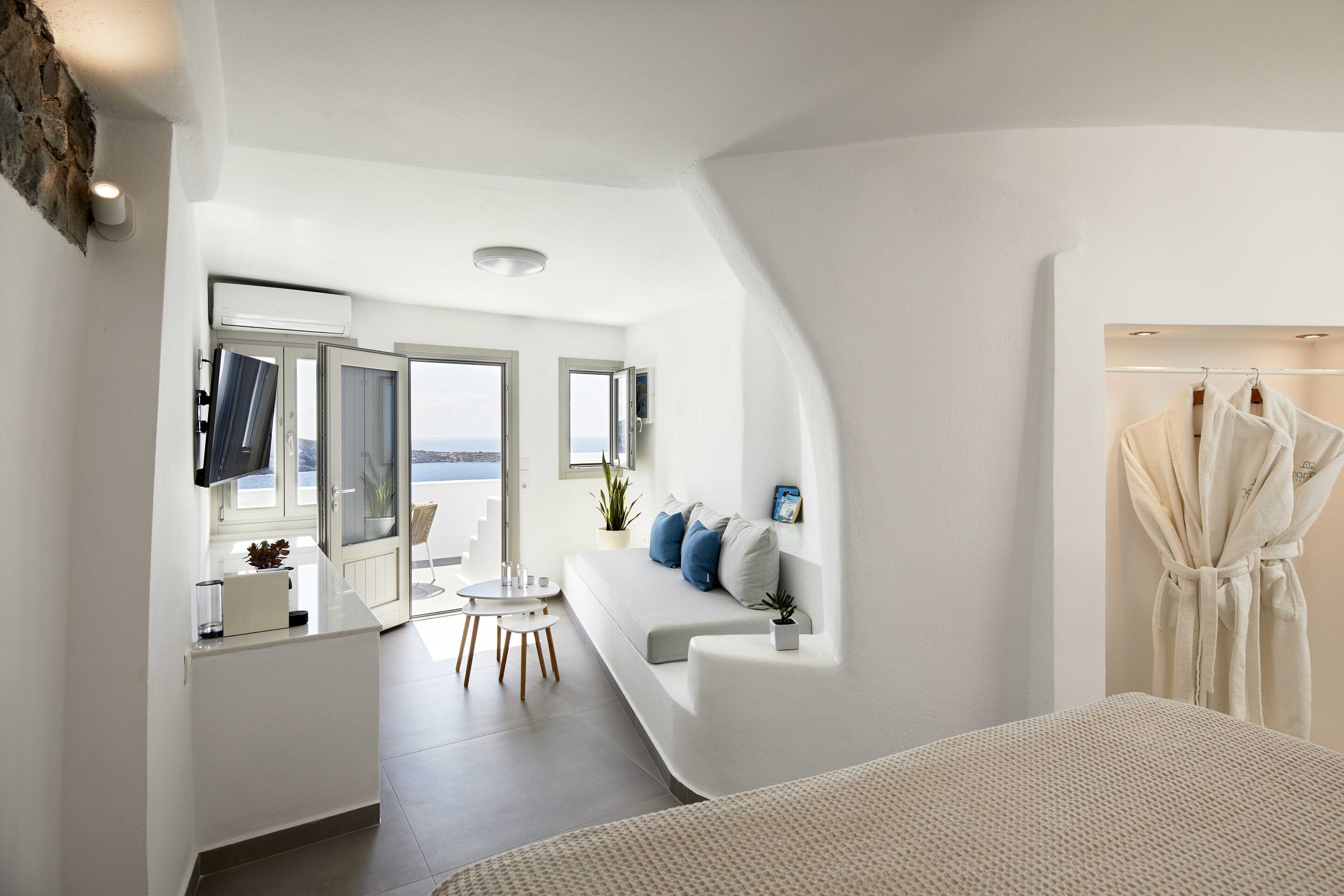 A modern, well-lit bedroom with a large bed, a seating area with a sofa and cushions, a small table, and a view of the outdoors through a sliding door.