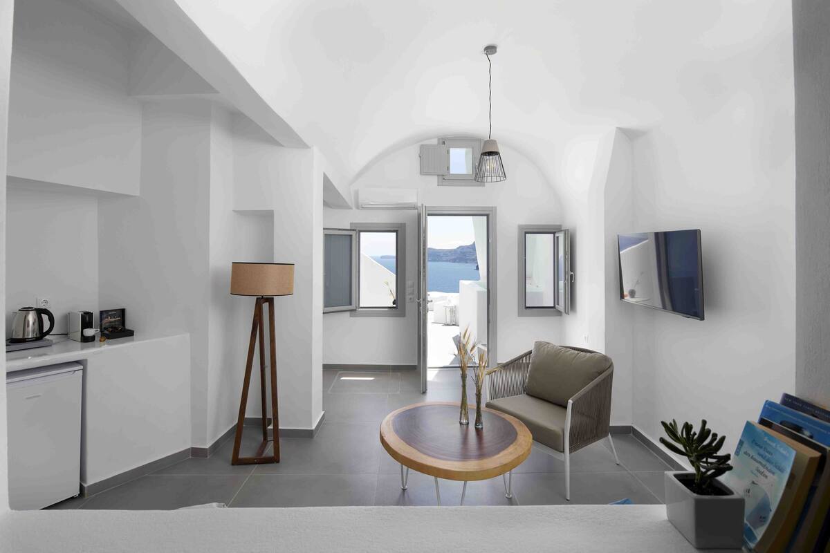 Een moderne, minimalistische woonkamer met een ronde houten salontafel, een bank, een televisie en uitzicht op zee.