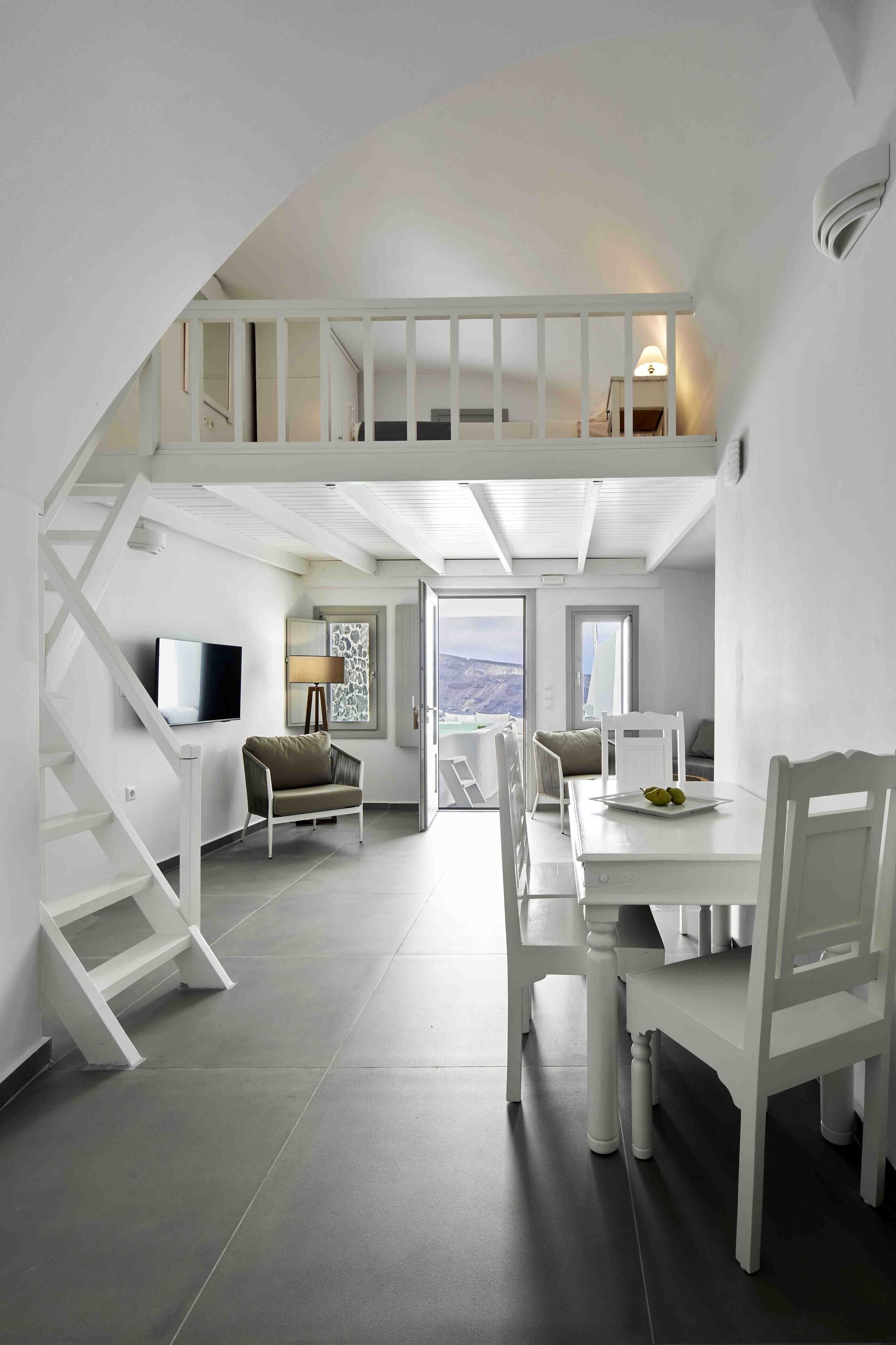 A spacious loft with a high ceiling, a staircase leading to an upper level, a dining area with a table and chairs, and a television mounted on the wall.