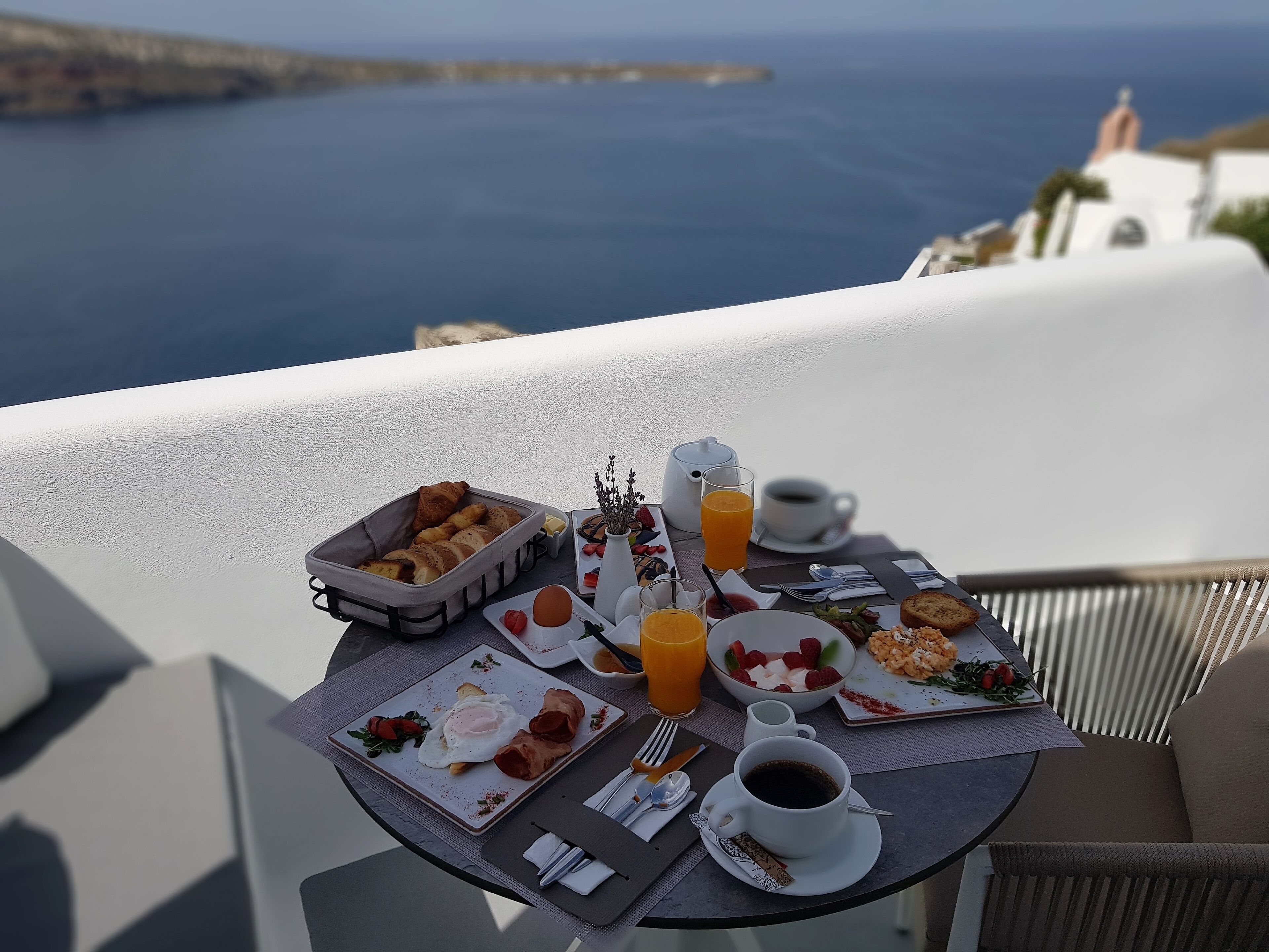 A breakfast table with various dishes, a coffee cup, and a glass of orange juice on a balcony overlooking the sea.