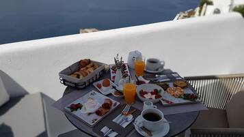 A breakfast table with various dishes, a coffee cup, and a glass of orange juice on a balcony overlooking the sea.