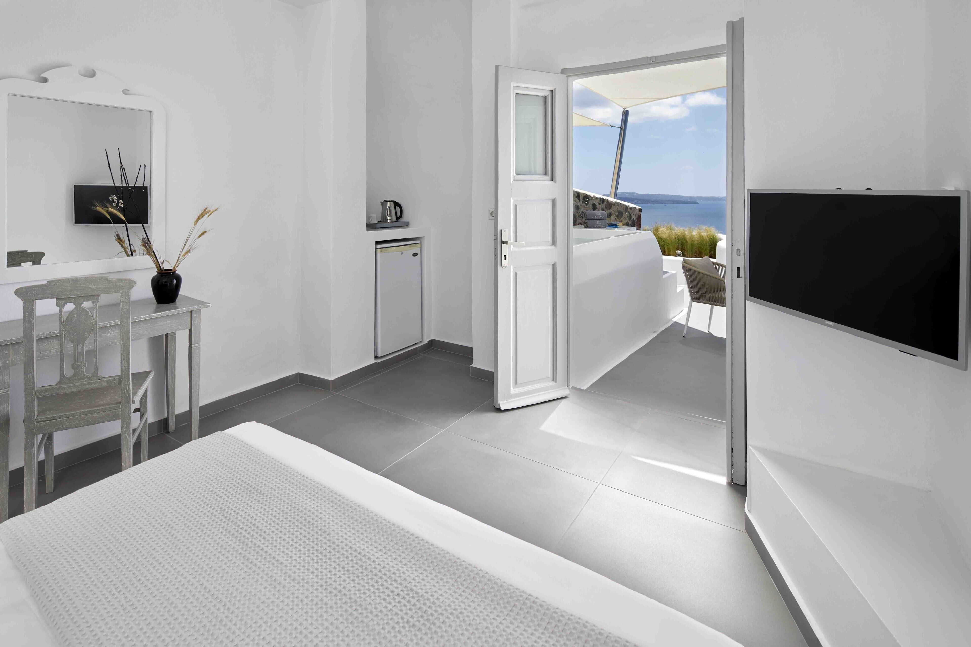 A modern hotel room with a bed, a desk, a small kitchenette, and a balcony with a view of the sea.