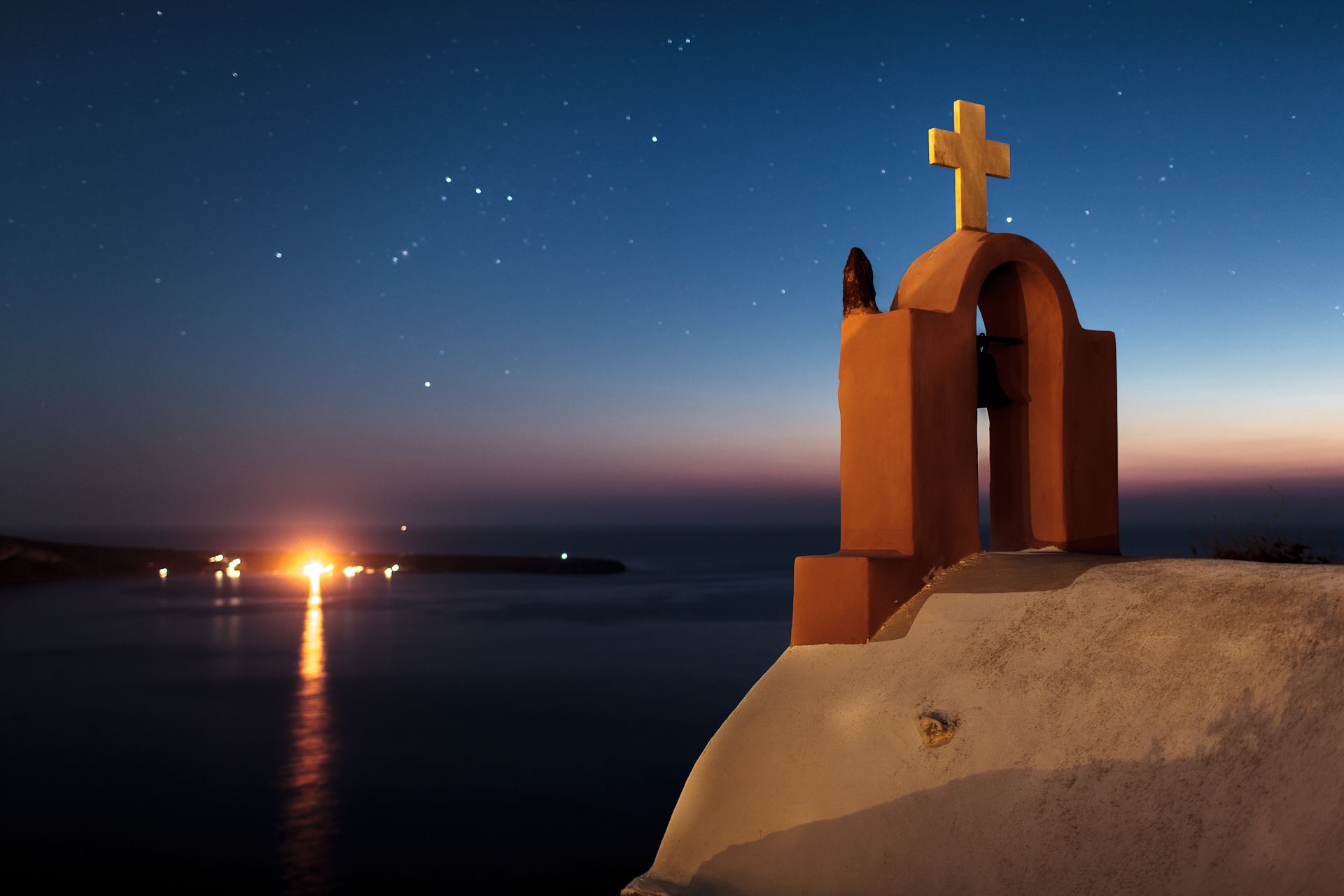 A coastal church with a cross on top, overlooking a calm sea and a city skyline at dusk.