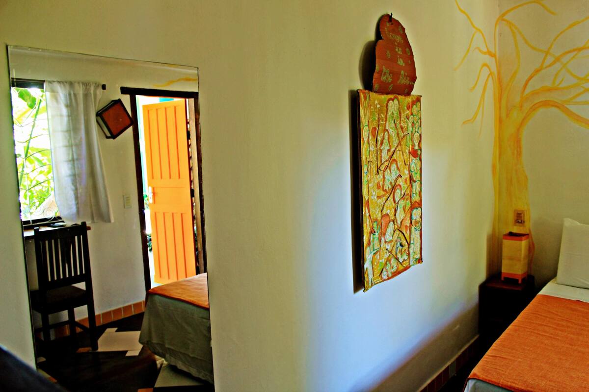 A room with a bed, a chair, a small table, and a wall with a tree painting.