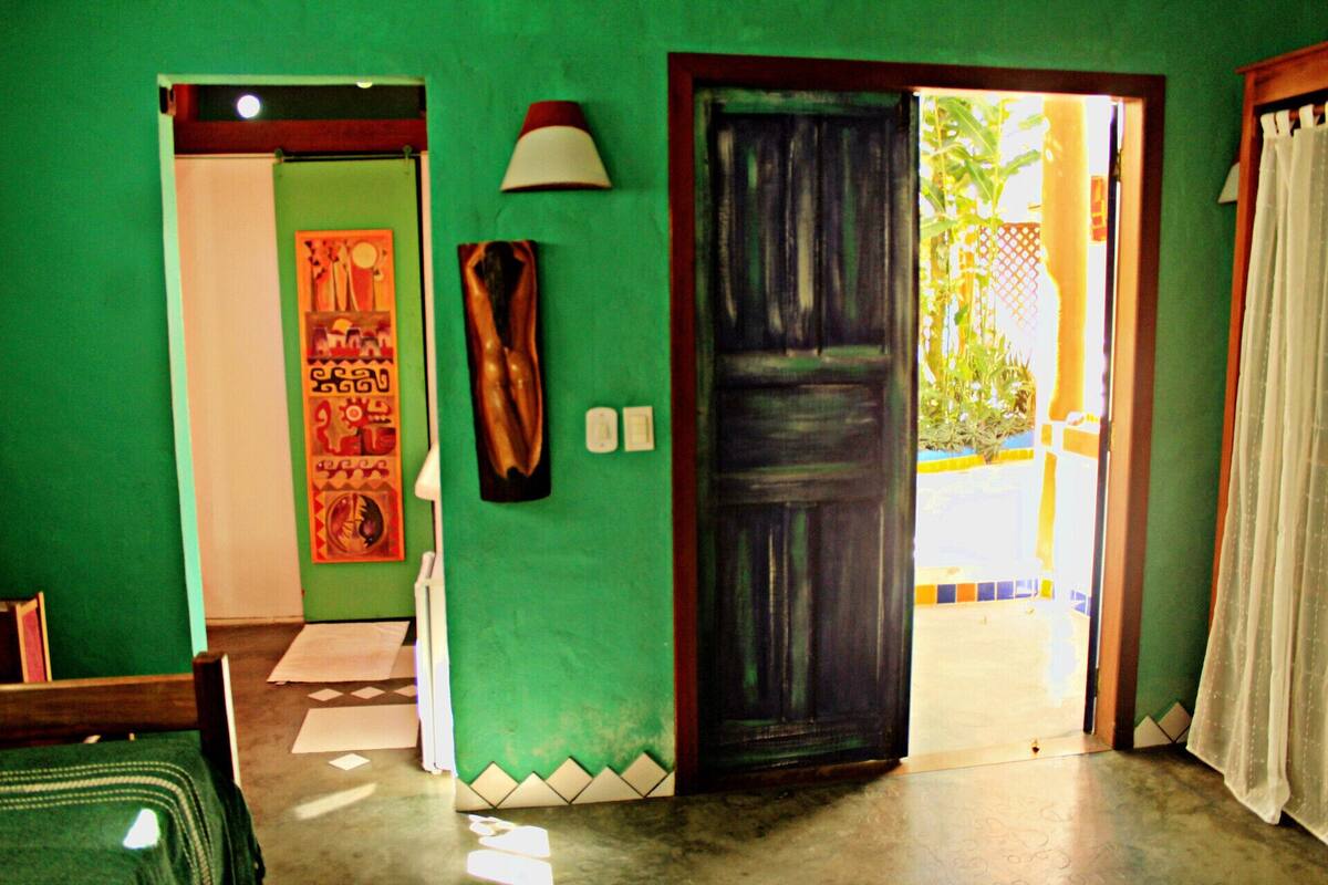 A room with a green wall, a wooden door, and a white lamp. There is a bed with a green blanket and a pillow.