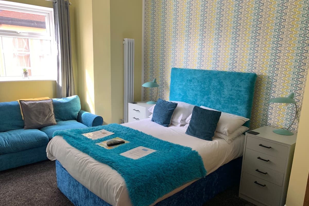 A bedroom with a large bed, a blue sofa, a bedside table with a lamp, and a radiator.