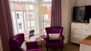 A room with two purple armchairs, a white dresser, a television, and a window with blinds.