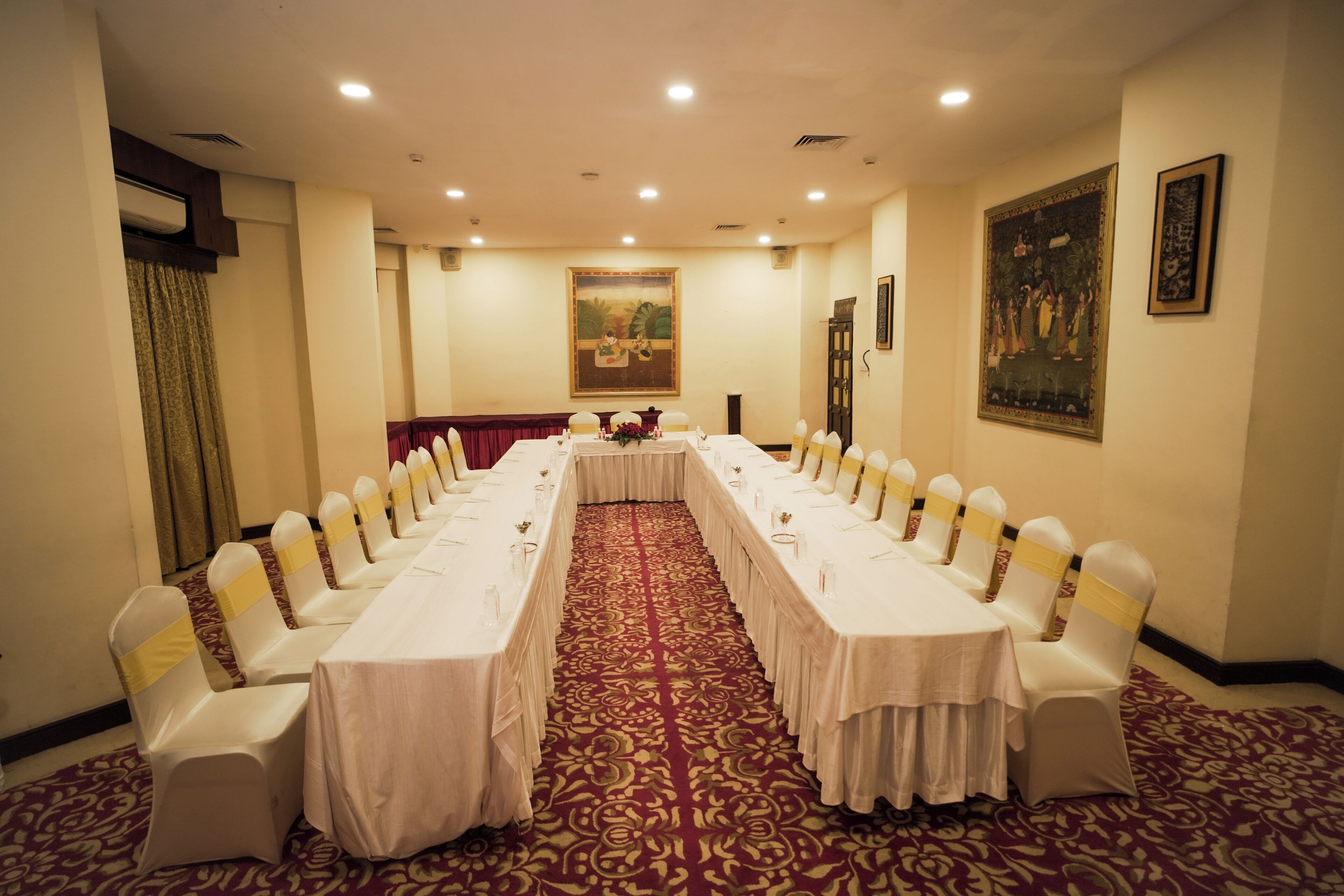 Un salón de conferencias con una larga mesa, sillas blancas adornadas con cintas amarillas, una alfombra roja y un cuadro en la pared.
