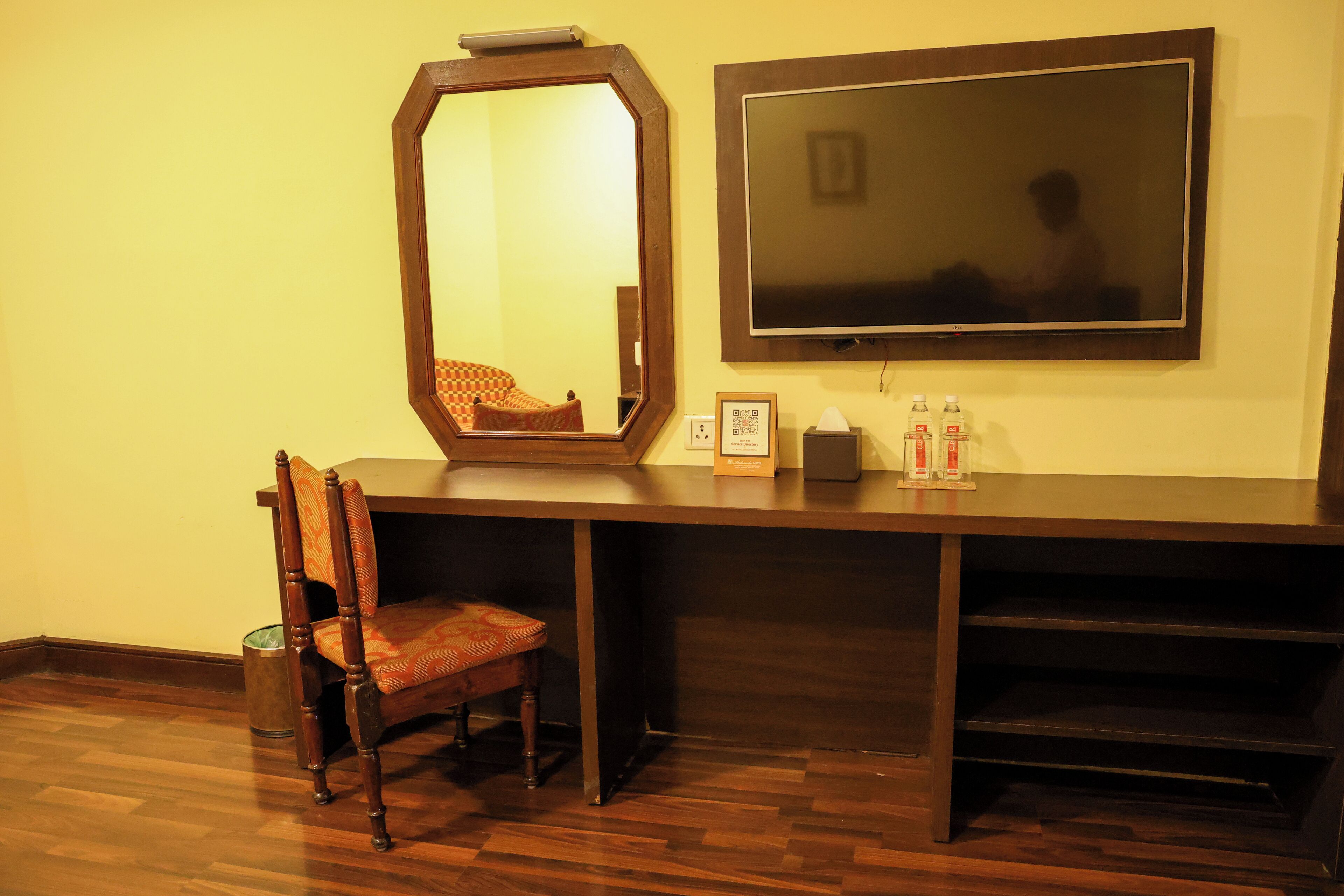 A room with a wooden desk, a chair, a large mirror, a television, and a trash can.