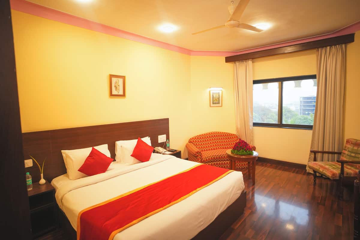 Habitación de hotel con una cama grande, una silla, una mesita con flores y una ventana con cortinas.
