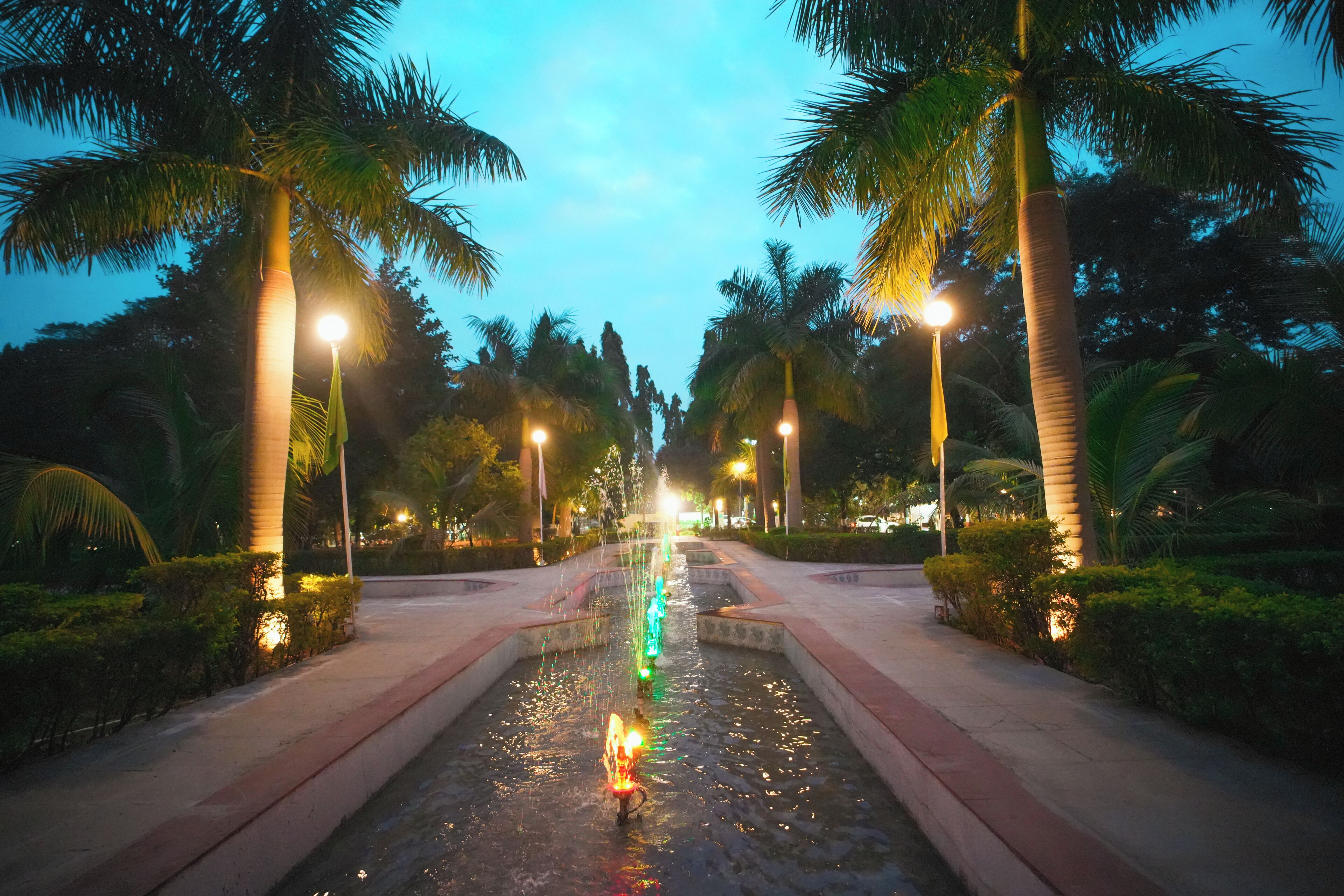 Une vue nocturne d’un élément décoratif aquatique illuminé, entouré de palmiers et bordé d’une allée pavée.