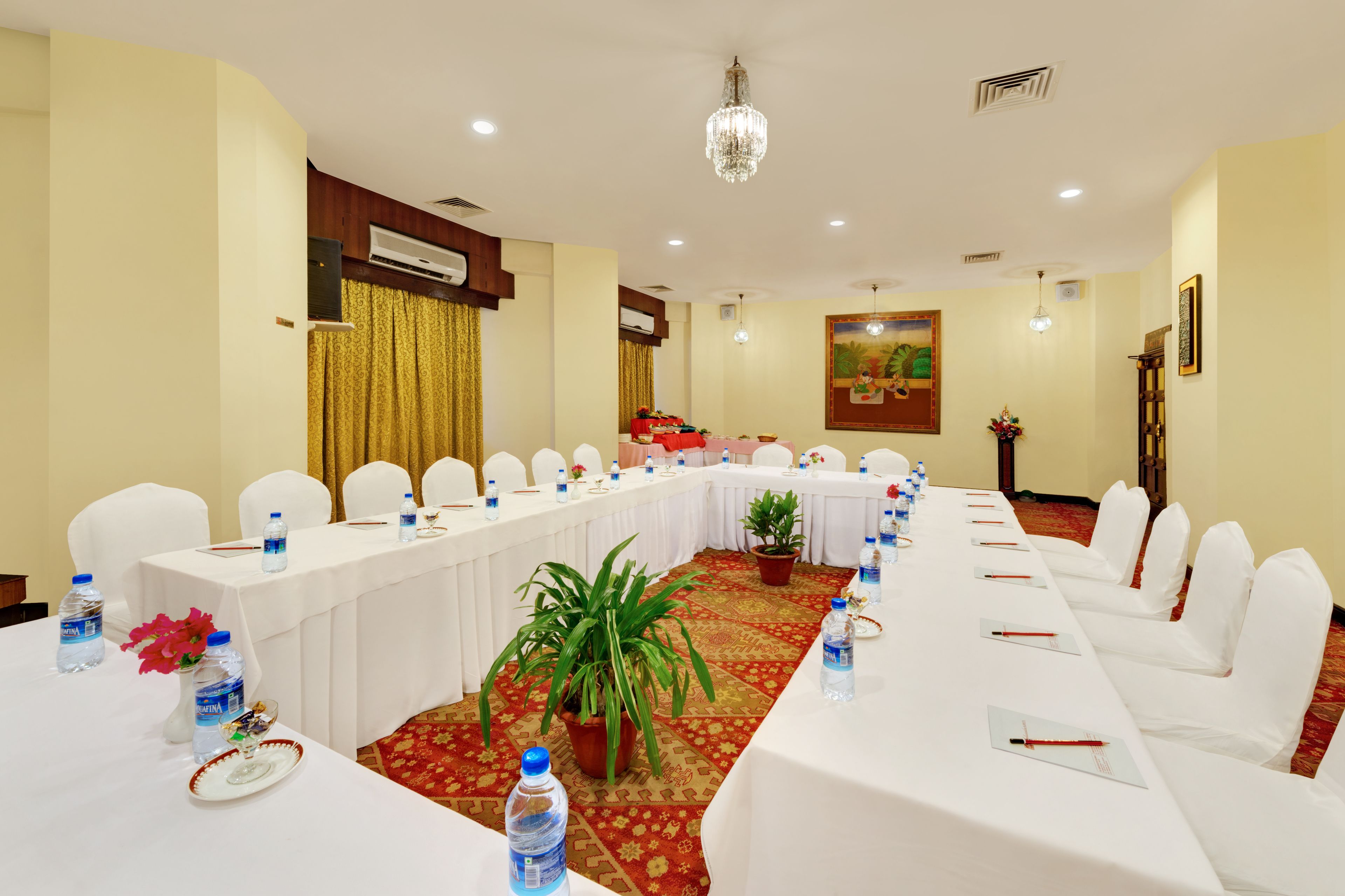 Un salón de conferencias con una larga mesa, sillas blancas, botellas de agua y una planta en maceta.