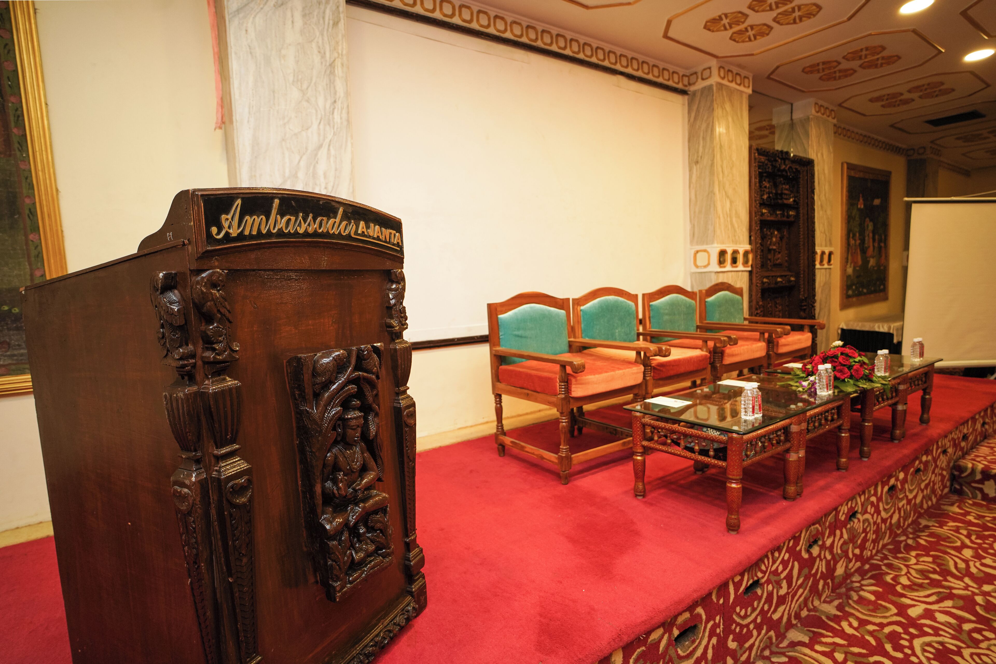 A conference room with a wooden podium, green upholstered chairs, a glass table, and a red carpet.
