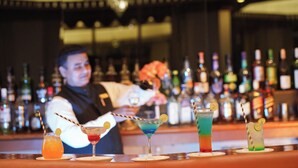 A bar with various cocktails on the counter, a bartender preparing drinks, and a background of neatly arranged liquor bottles. - Ambassador Ajanta (Chhatrapati Sambhajinagar)