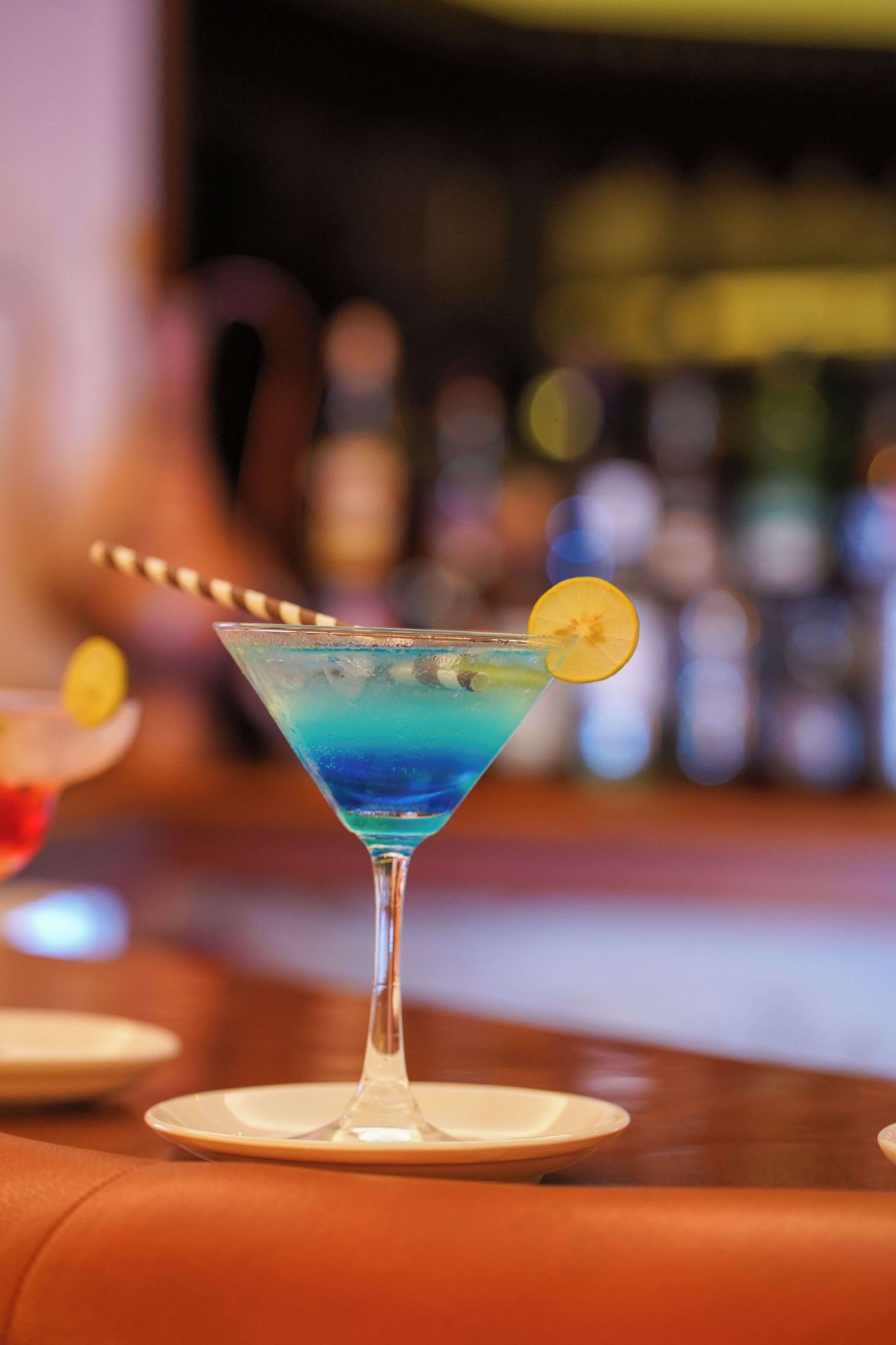 A martini glass with a blue liquid and a lemon slice garnish on a bar counter.