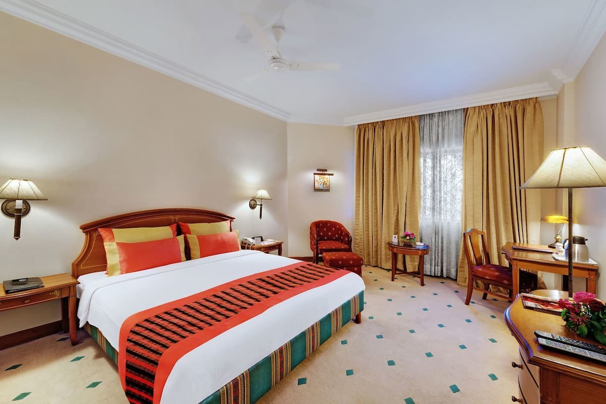 Una habitación de hotel con cama, dos sillas, un escritorio, una lámpara, teléfono y ventana con cortinas.